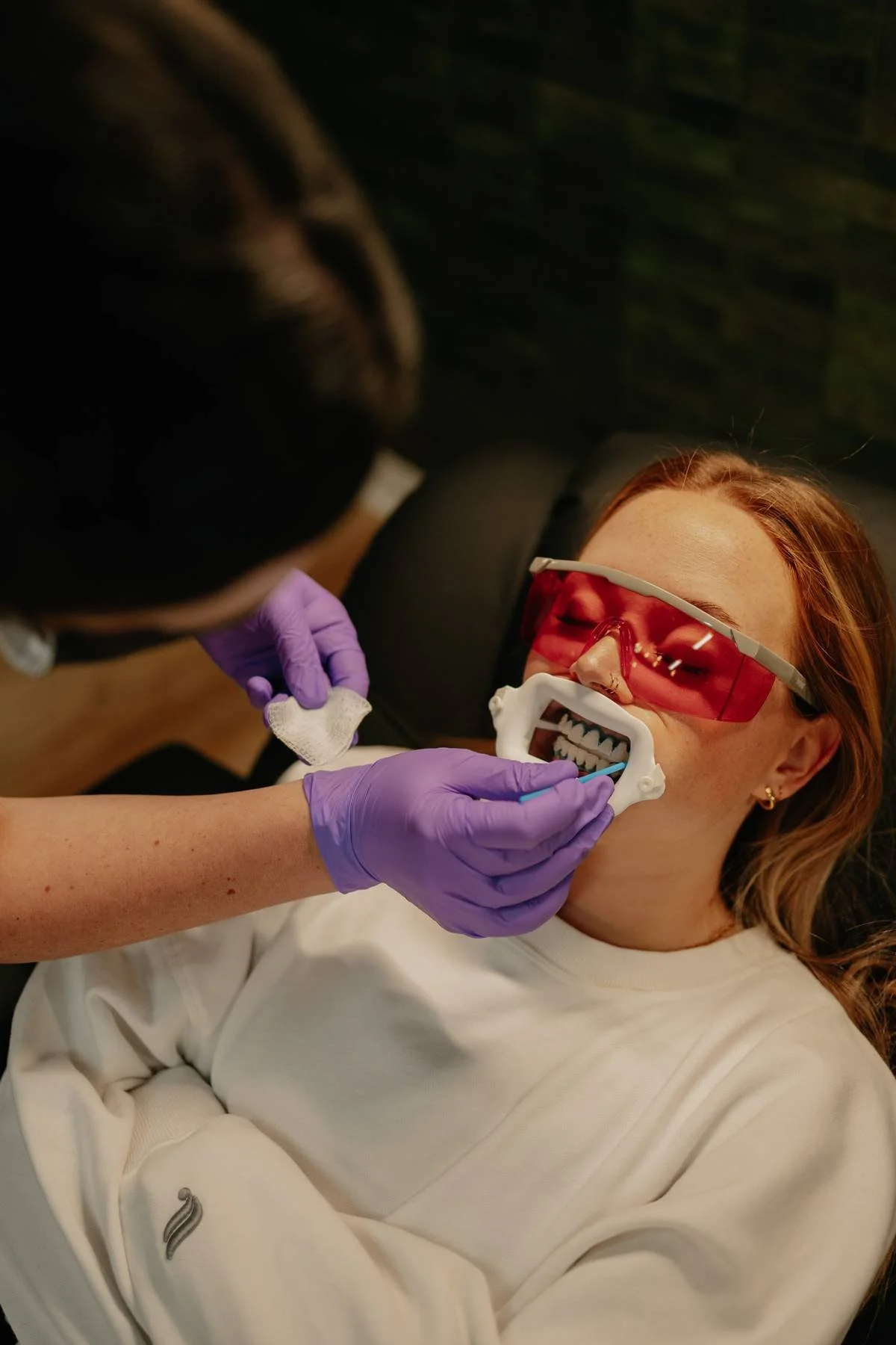 A woman with red hair wearing protective pink-tinted glasses is lying back during a dental exam. A dental professional, wearing purple gloves, is holding a dental mirror and a gauze pad near her mouth, which has a dental retractor in place. The woman appears relaxed during the procedure.