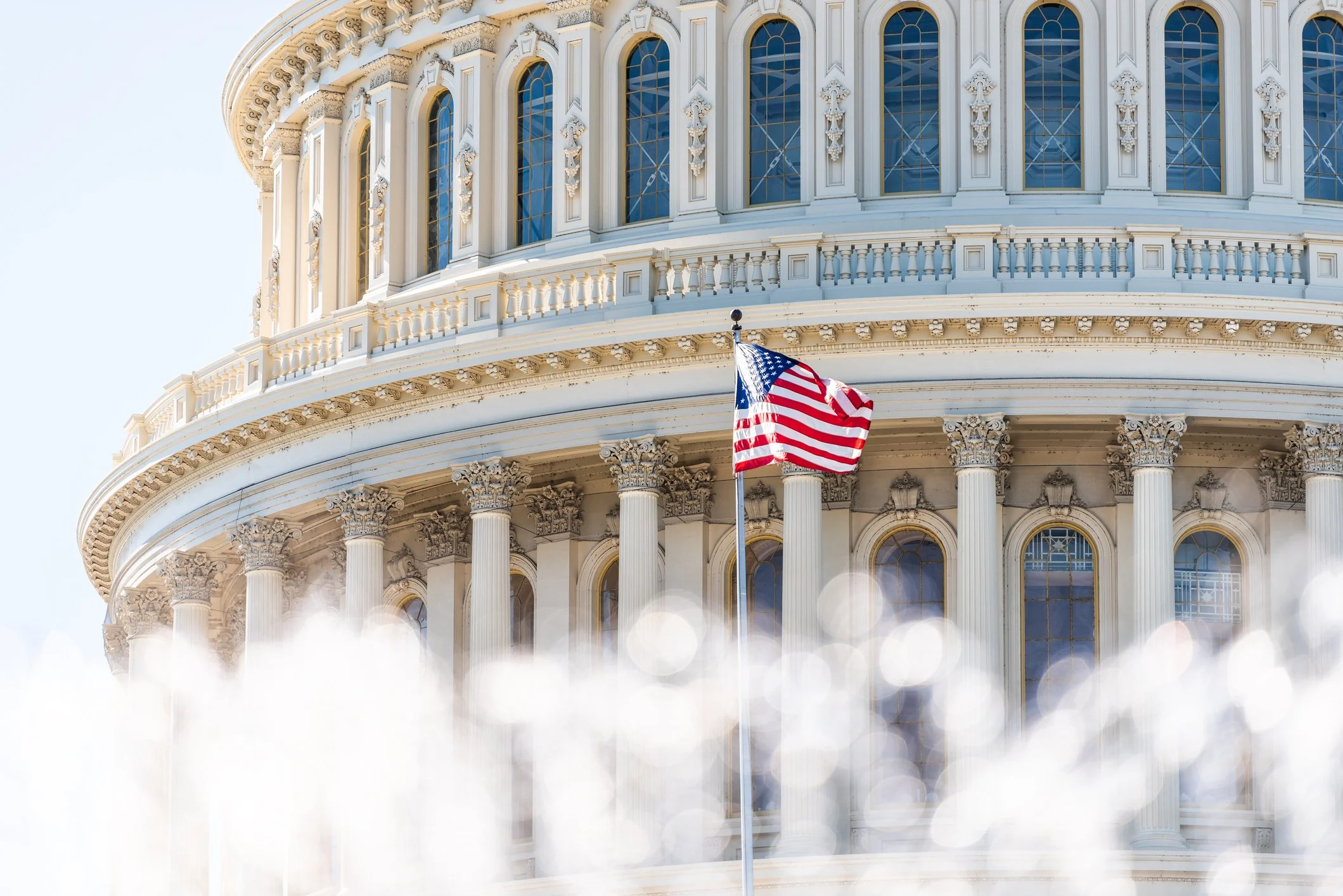 Part of the United States Capitol building with an American flag in front, showing ornate columns and windows.