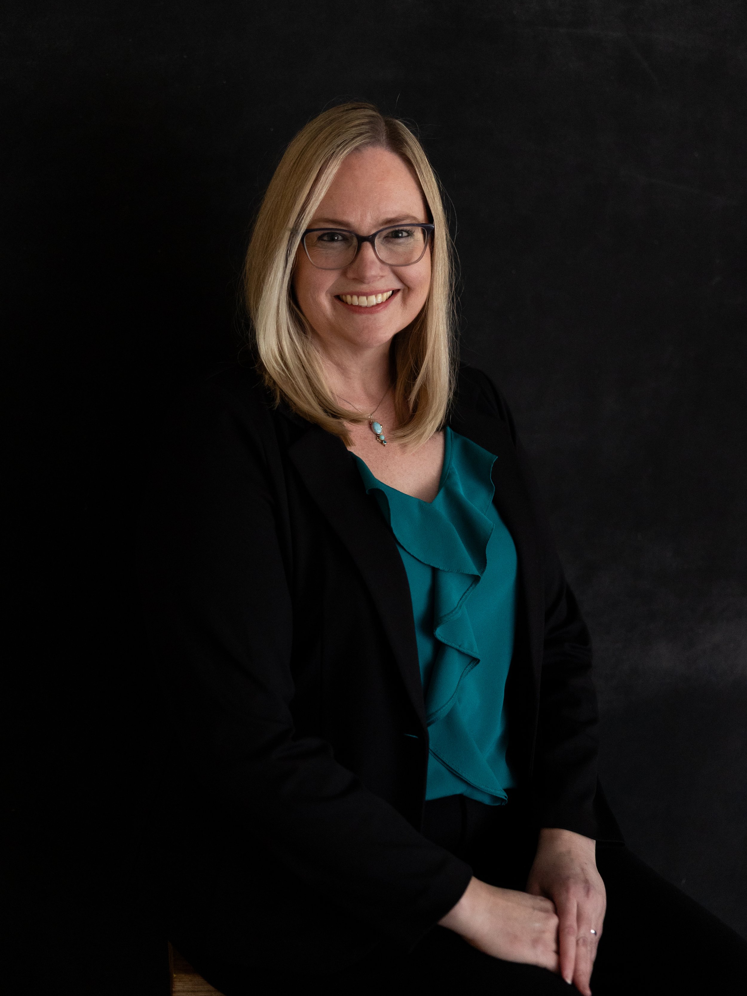 A woman with blonde hair, glasses, and a smile, wearing a black blazer and teal blouse, sitting against a dark background.