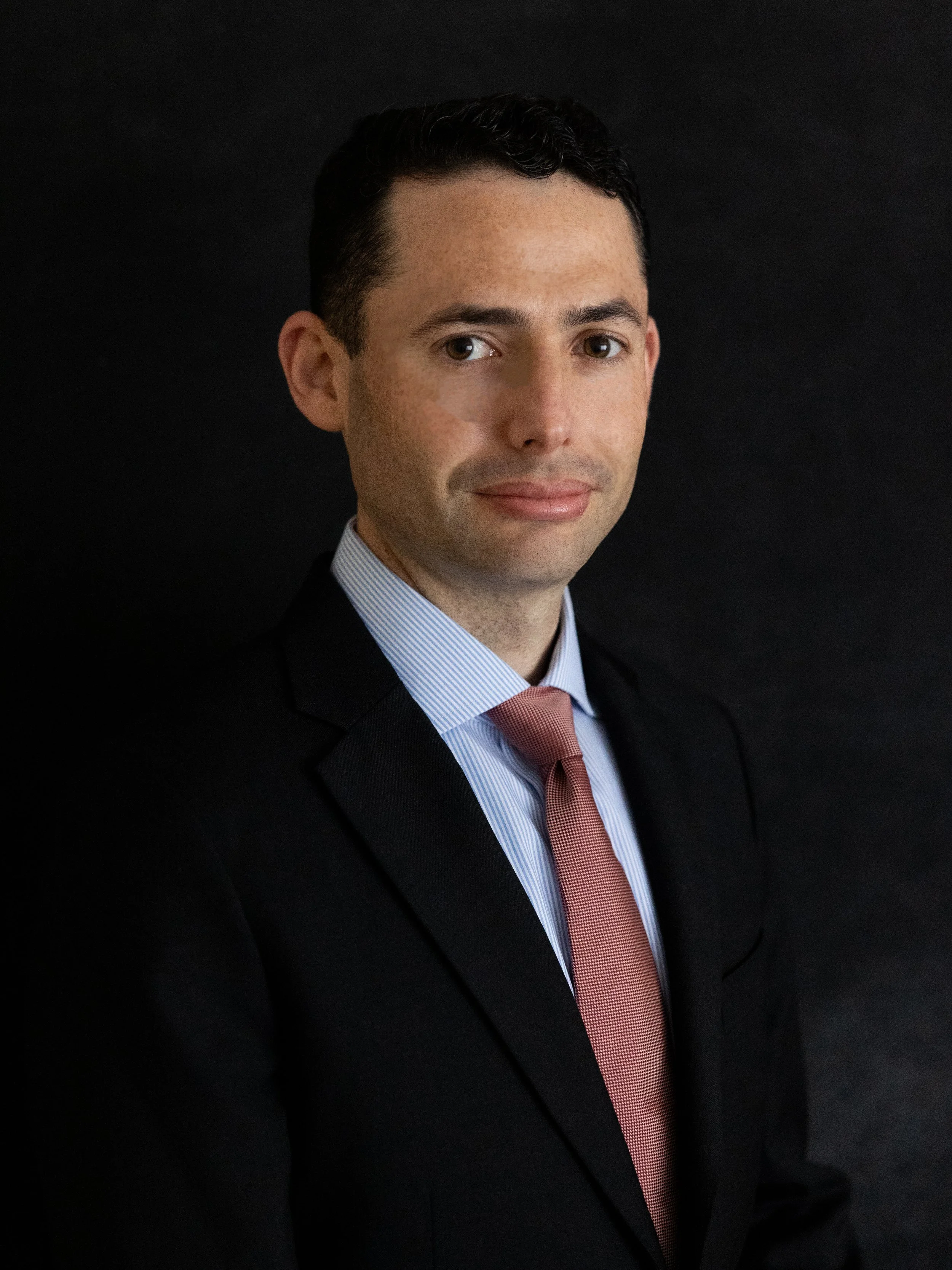 Professional headshot of a man in a suit and tie against a dark background.