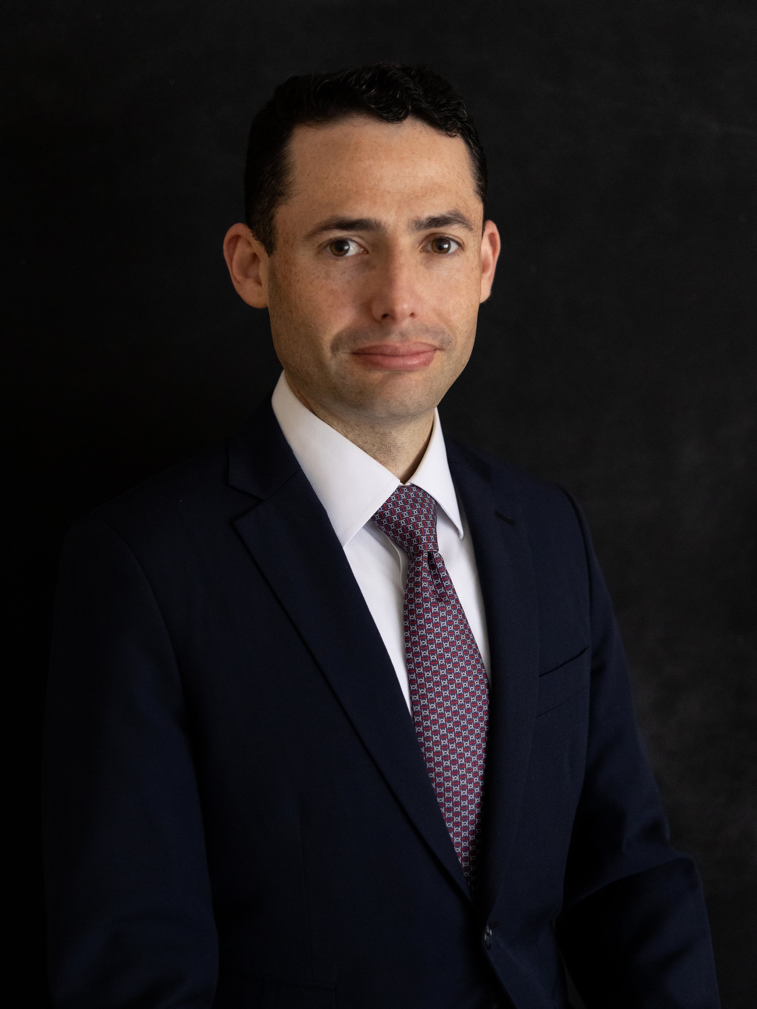 Formal portrait of a man with dark hair, wearing a navy suit, white shirt, and patterned tie, against a black background.