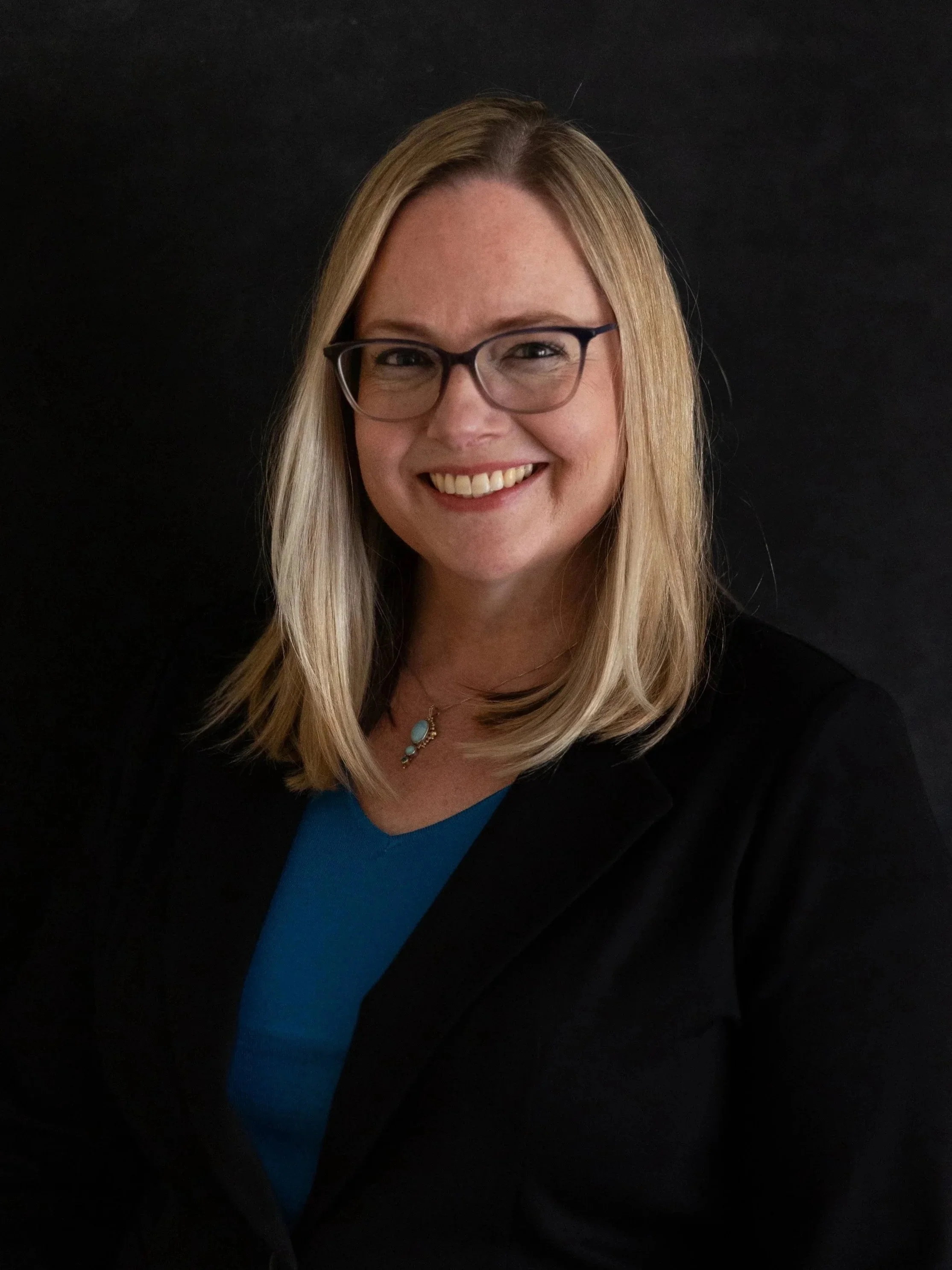 A woman with shoulder-length blonde hair, wearing glasses, a black blazer, and a blue top, smiling against a dark background.
