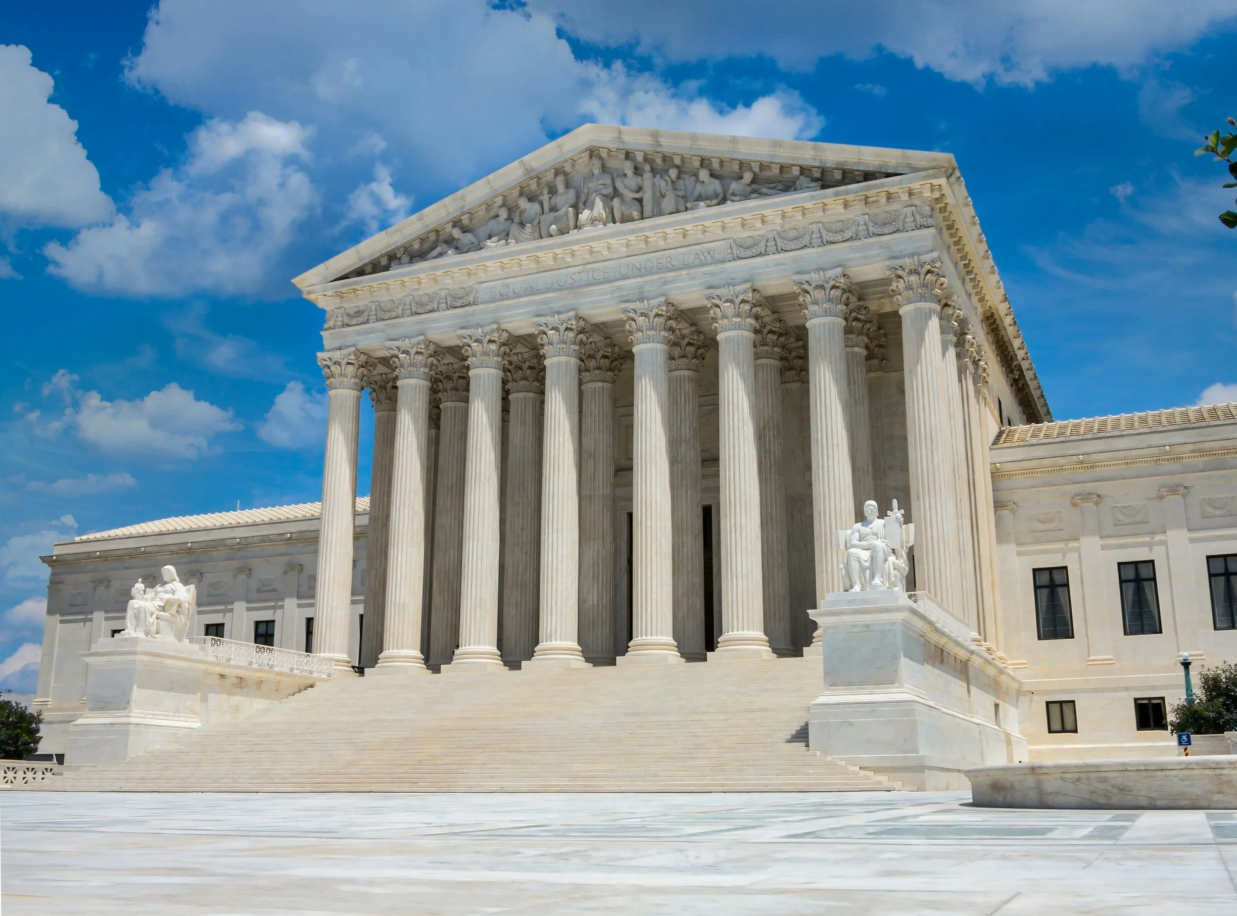 The United States Supreme Court building with grand columns, statues, and steps, under a partly cloudy blue sky.