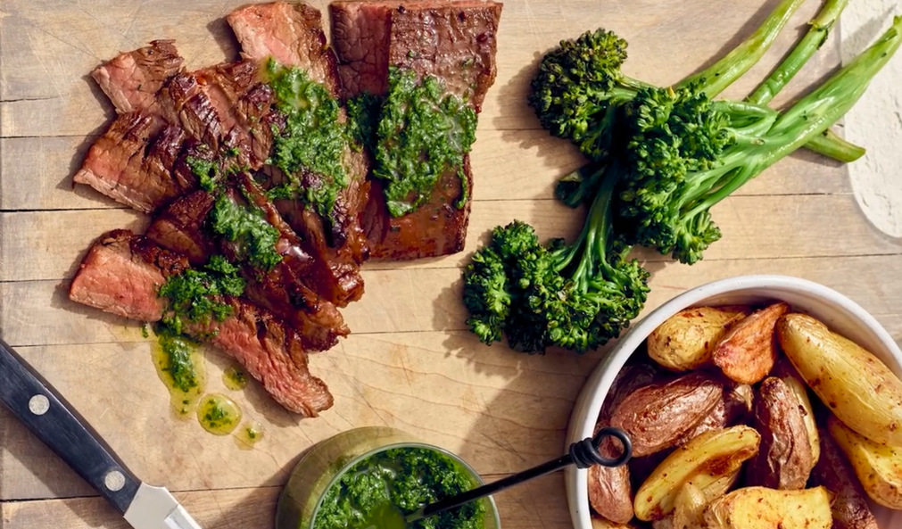 Cooked steak slices topped with green herb butter, accompanied by steamed broccoli, roasted potatoes, and a jar of herb sauce on a wooden cutting board.