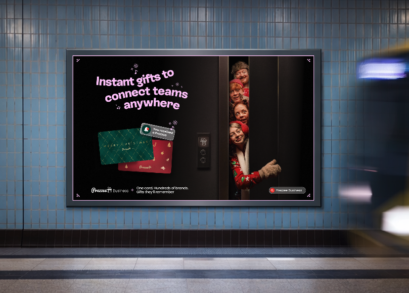 Digital advertisement in a subway station for a gift card service, showing a virtual elevator with four people's faces peeking out, and colorful cards with the text "Instant gifts to connect teams anywhere".