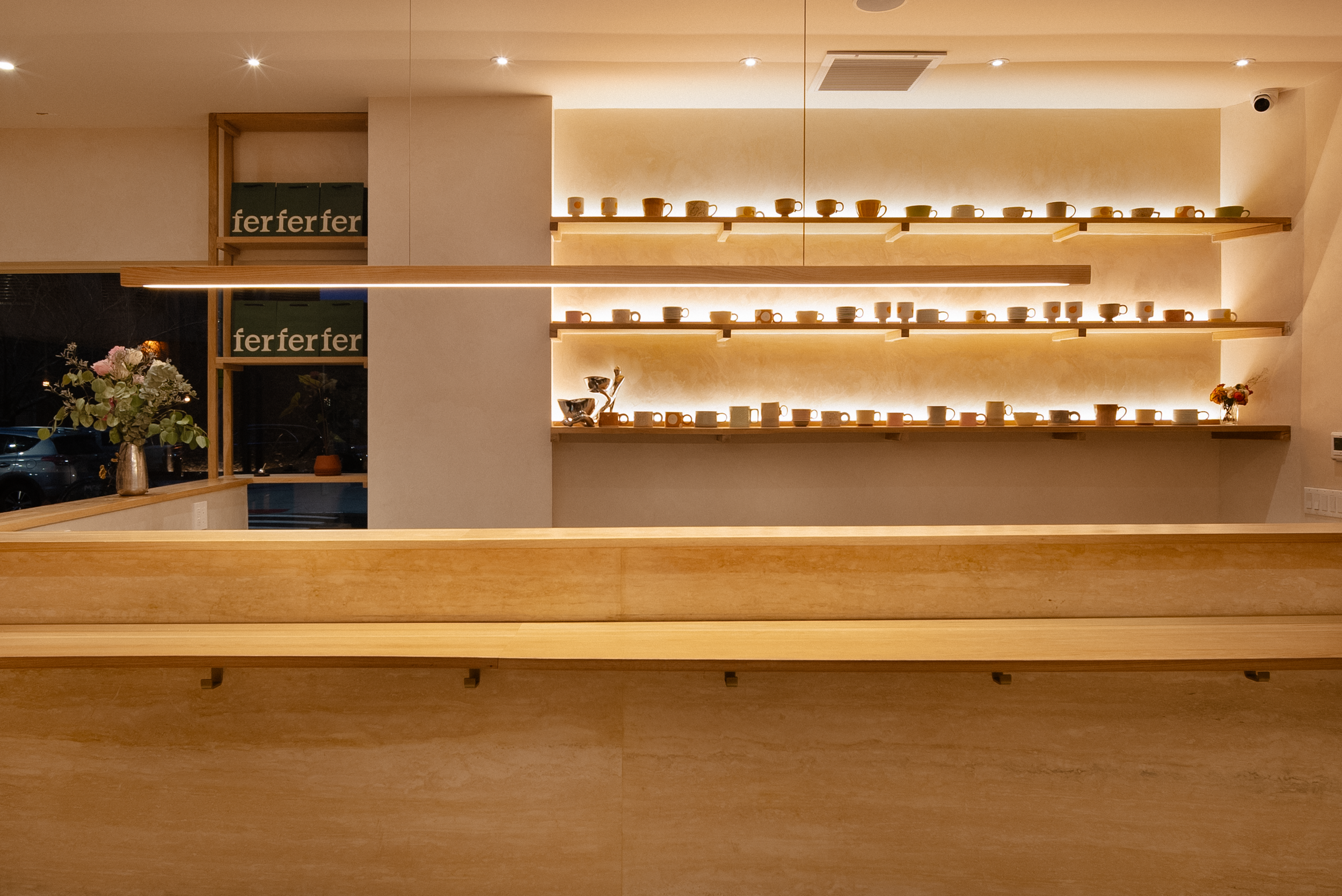 Interior of a minimalist cafe or coffee shop with wooden shelves displaying cups, a flower arrangement on the counter, and warm lighting.