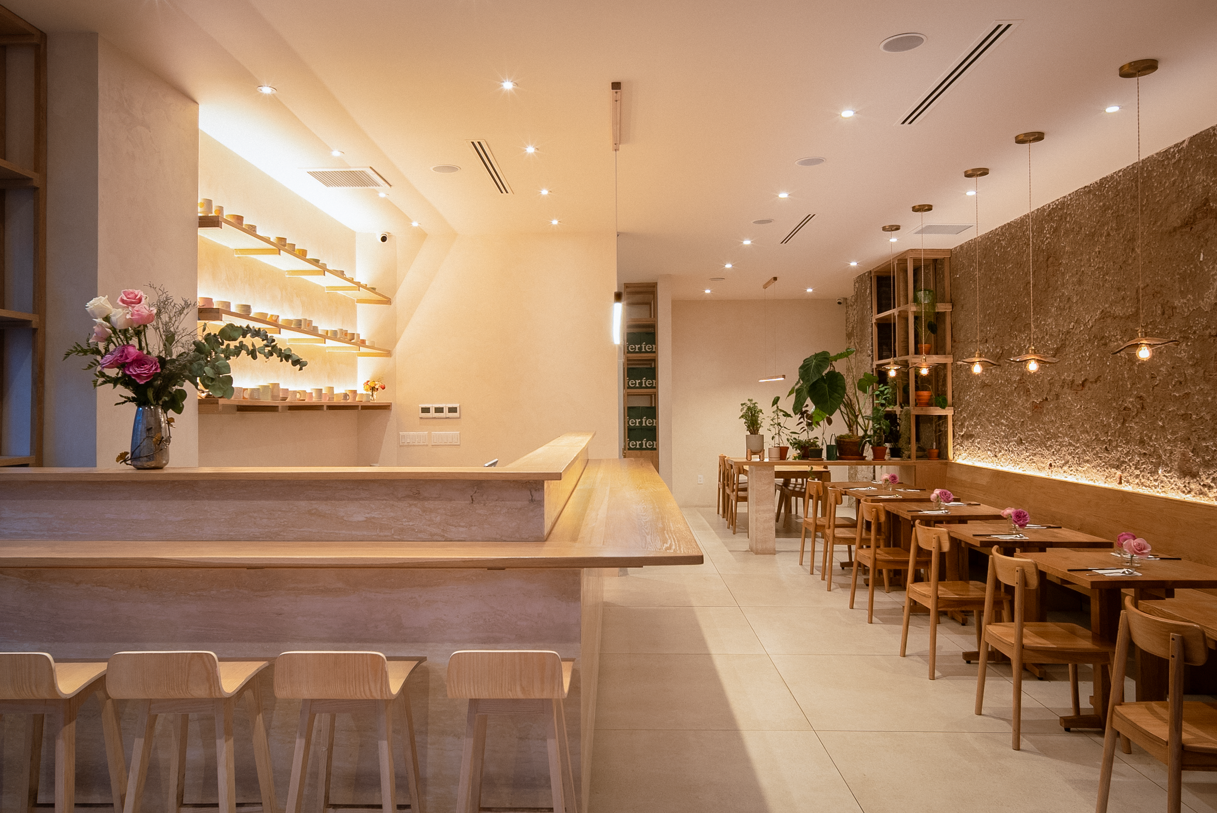 Interior of a modern restaurant with a long wooden counter, flower arrangement, and a seating area with small tables and chairs, decorated with pink flowers and green plants.