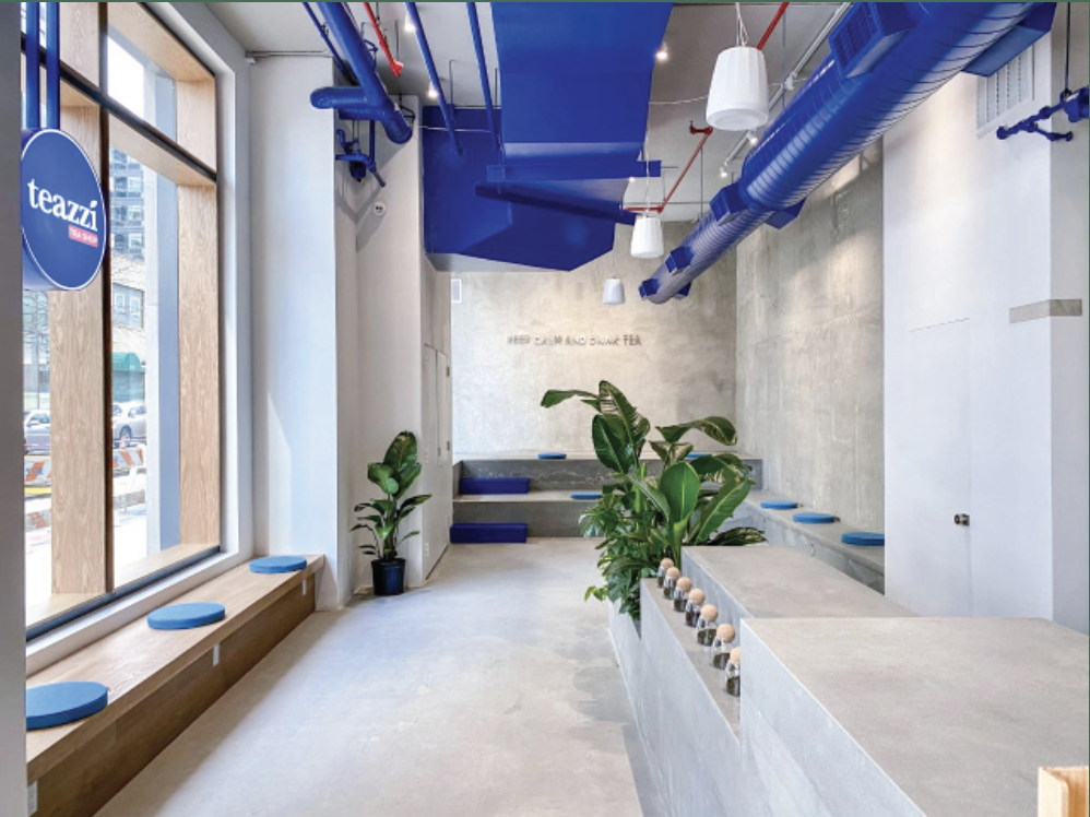 Modern tea shop interior with large windows, blue and white decor, plants, seating area with blue cushions, and a sign that reads 'HERBAL & SUPER BLEND TEA'.