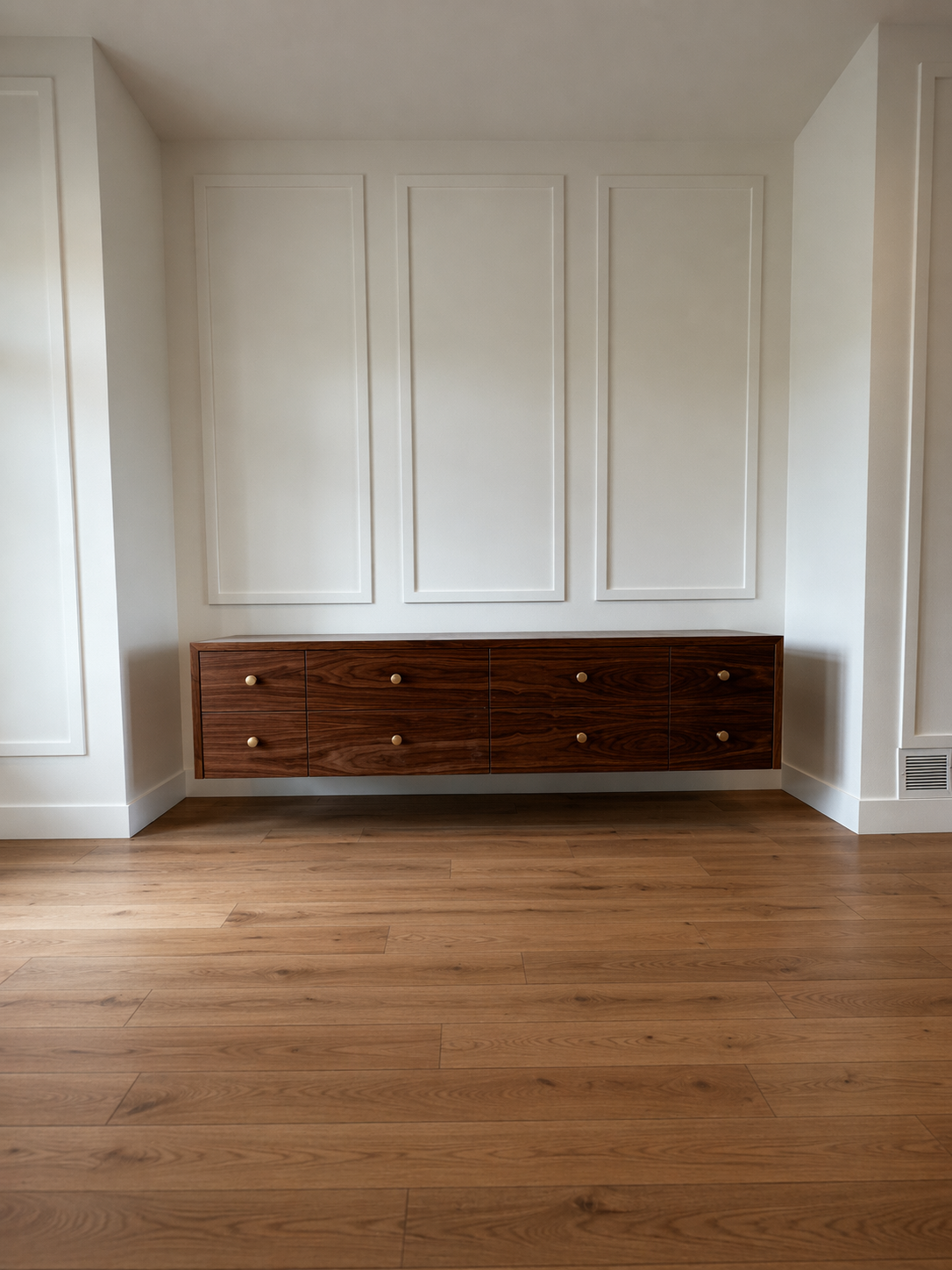 Empty room with white walls and wooden flooring, featuring a wooden dresser with six drawers and round knobs against the wall.