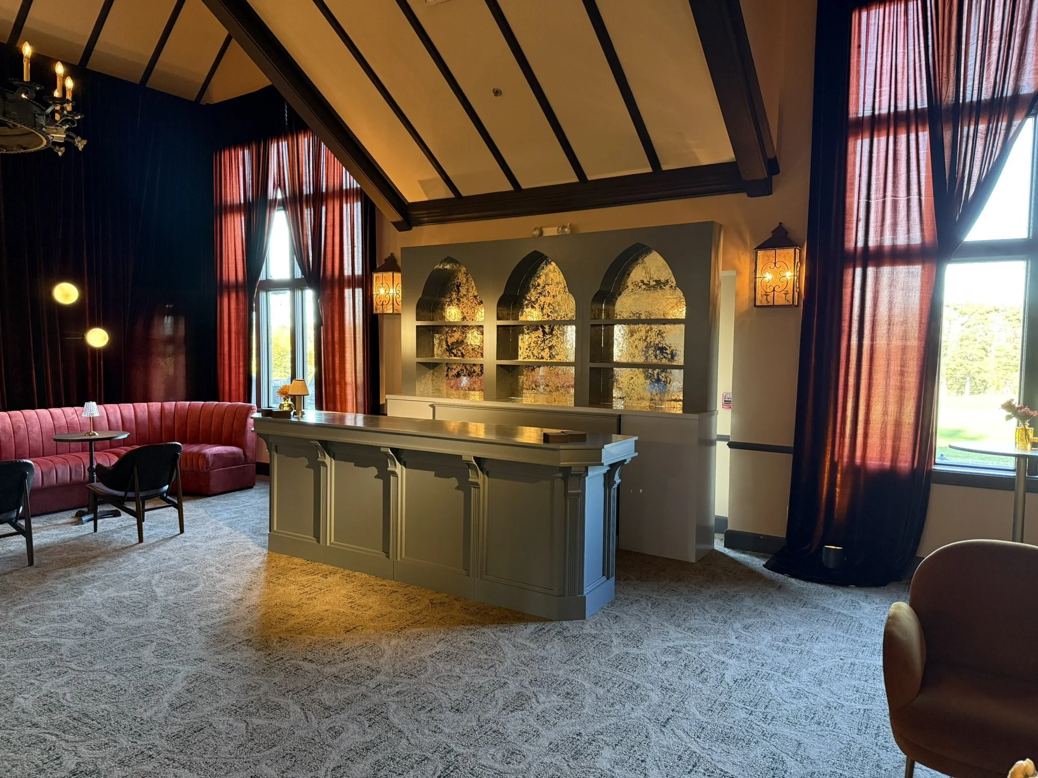 A cozy lounge area with a red velvet curved sofa, black chairs, small side tables with lamps, a bar counter, and large windows with dark curtains, illuminated by natural daylight.