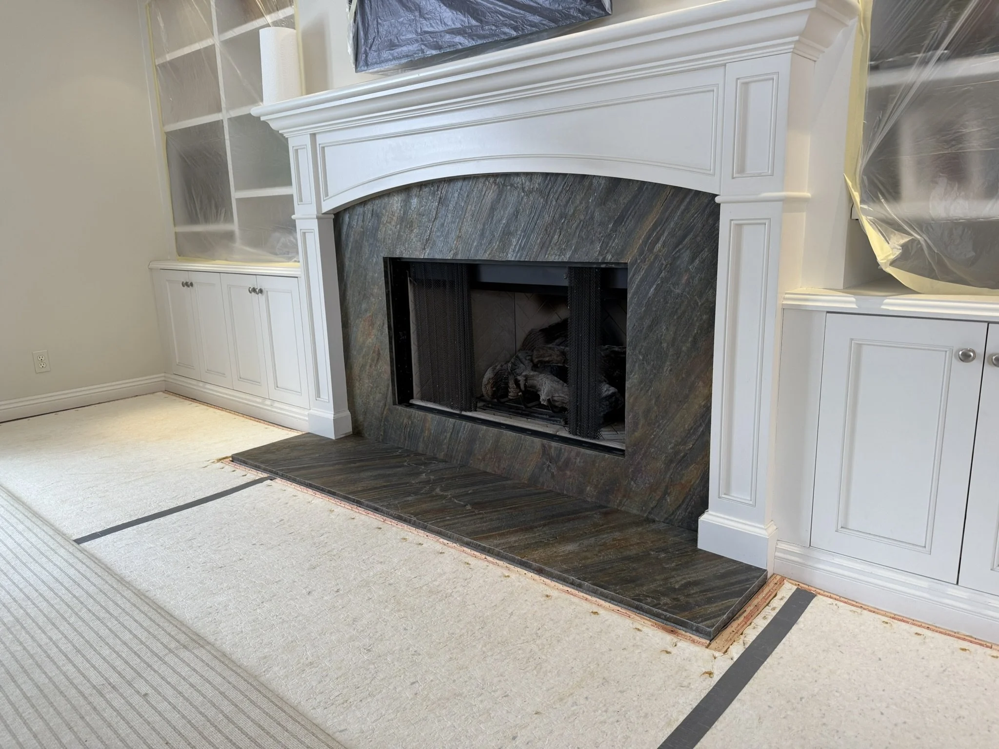 Living room fireplace with a white mantel and a dark stone surround, with construction materials and plastic coverings nearby.