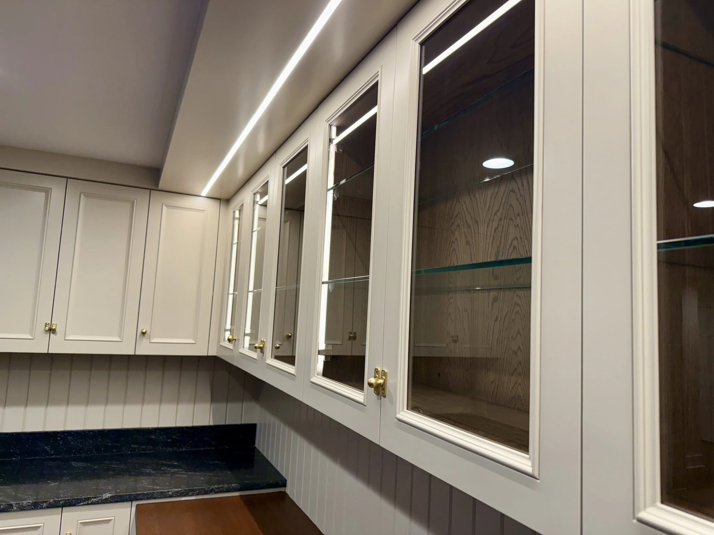Kitchen cabinets with glass doors, white and wood finish, under LED lighting, with a black countertop and a wooden cutting board.