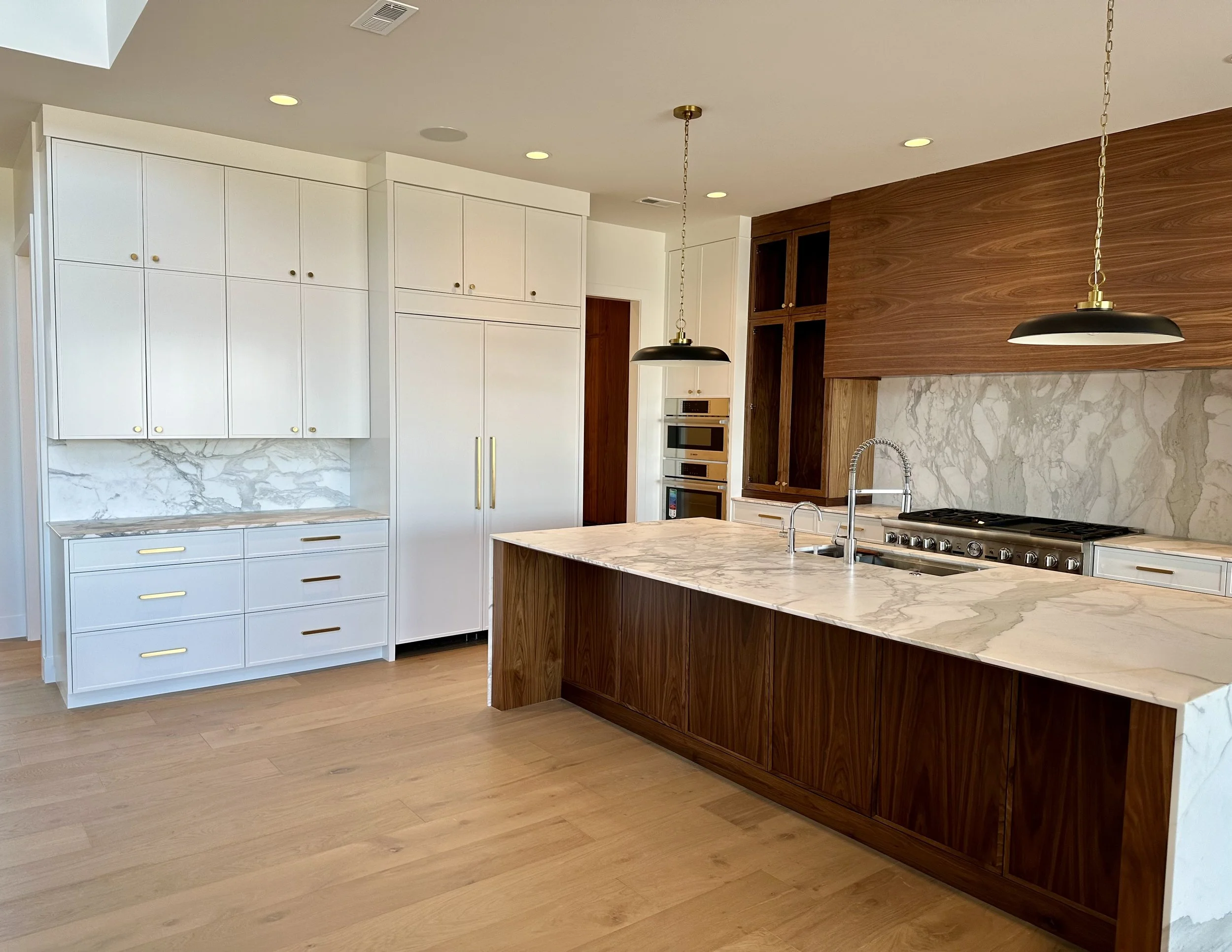 Modern kitchen with white and wooden cabinetry, marble countertops, and a large island with a sink and gold hardware, pendant lights hanging above.