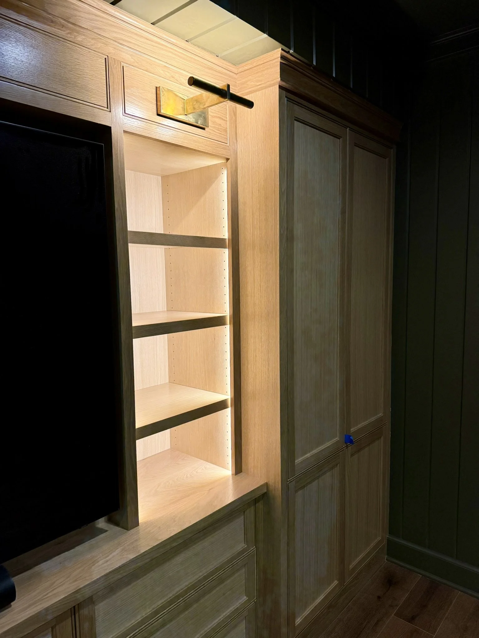 Wooden cabinets and open shelving unit with interior lighting, next to a dark plain wall and a wooden floor.