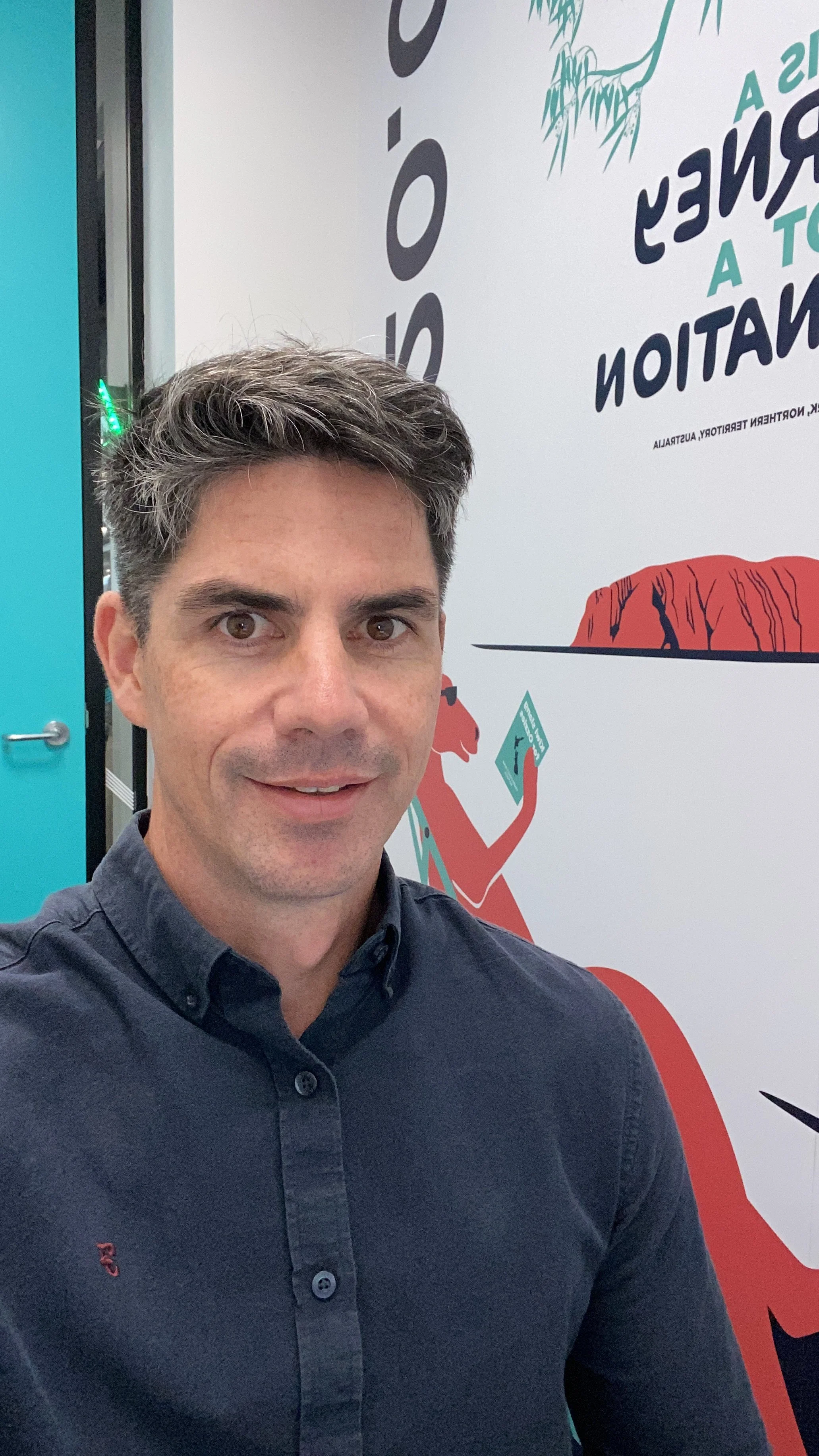 Selfie of Warren Hills in a navy blue shirt standing in front of a colorful wall poster with a cartoon bear holding a map.