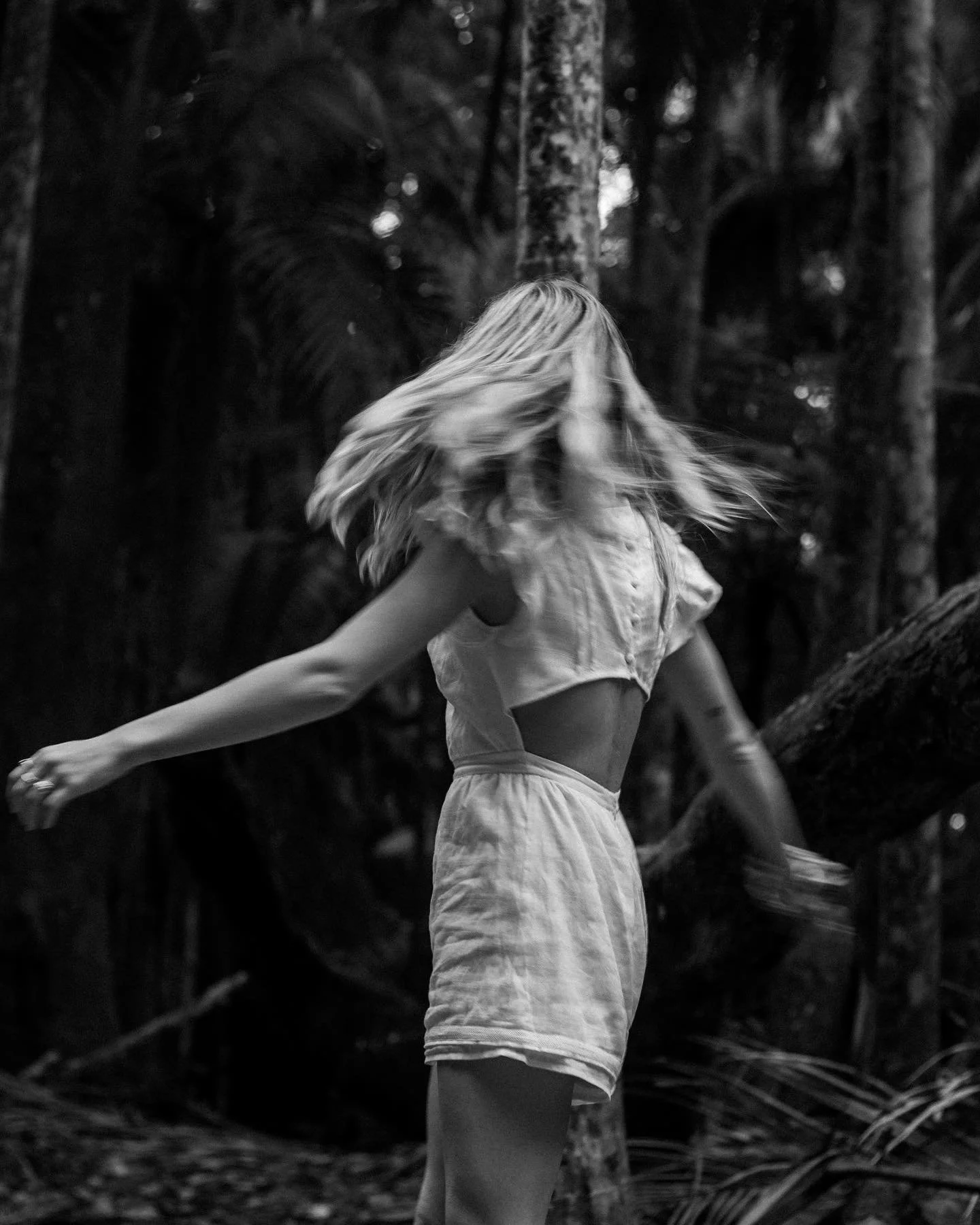A woman with long, flowing hair dancing in a forest, wearing a light-colored top and shorts, captured in motion.