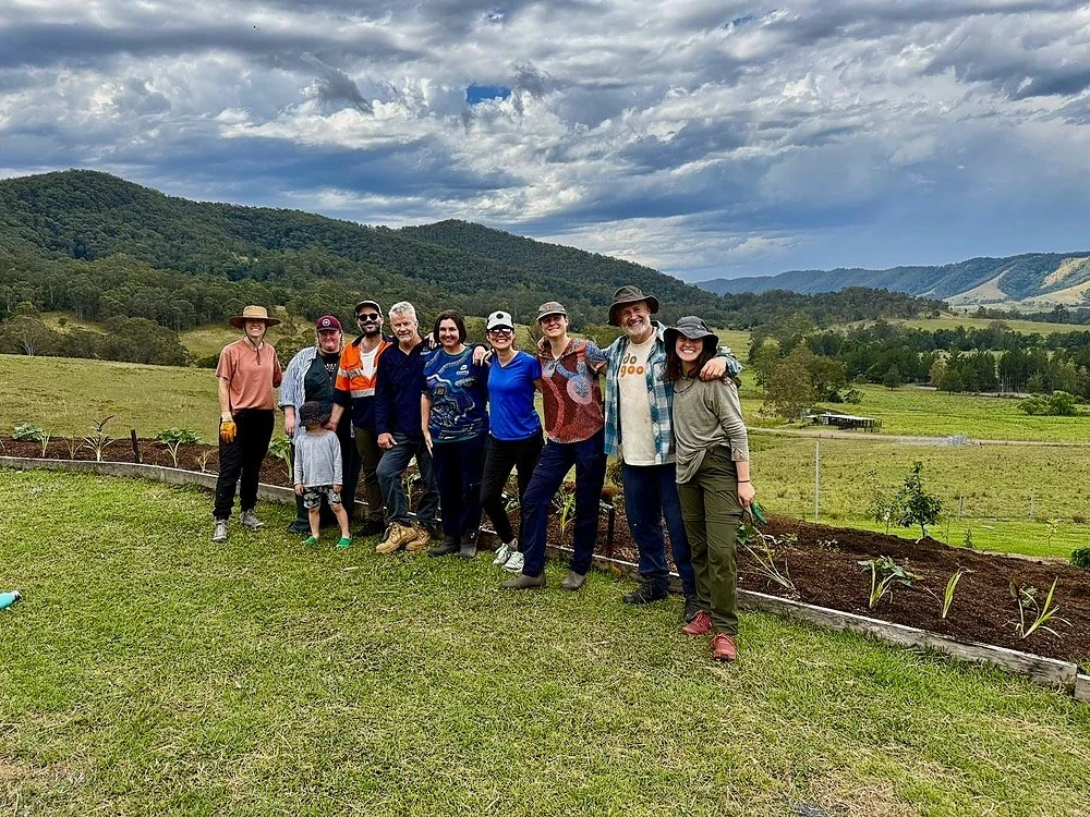Group of people from one of Djanaba Farm's intensives standing outdoors on a grassy area, with a backdrop of rolling green hills and a cloudy sky, during daytime.