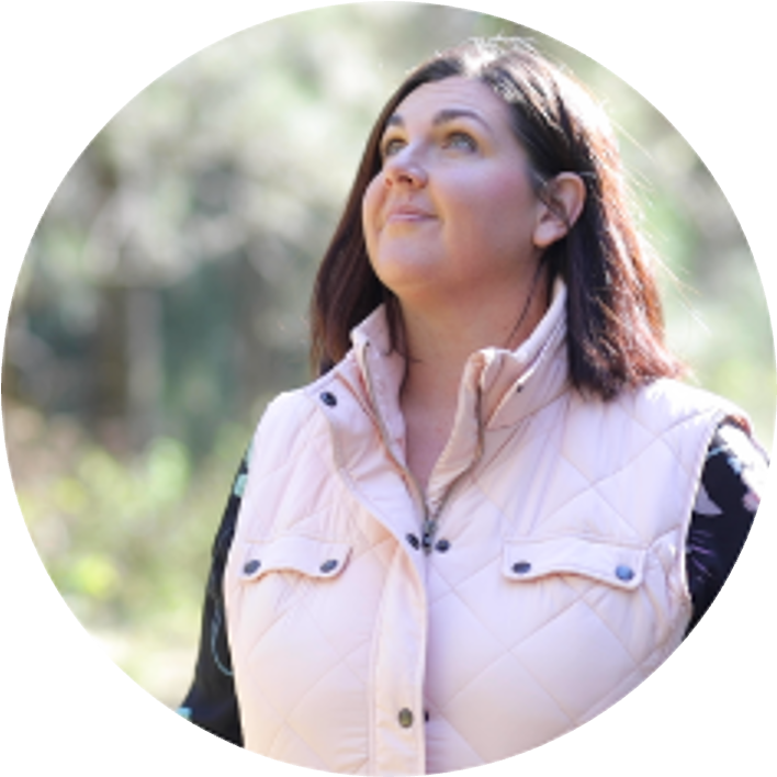 Jocelyn King, a woman with brown hair looking upwards outdoors in natural light, wearing a light pink quilted vest over a black and white shirt.