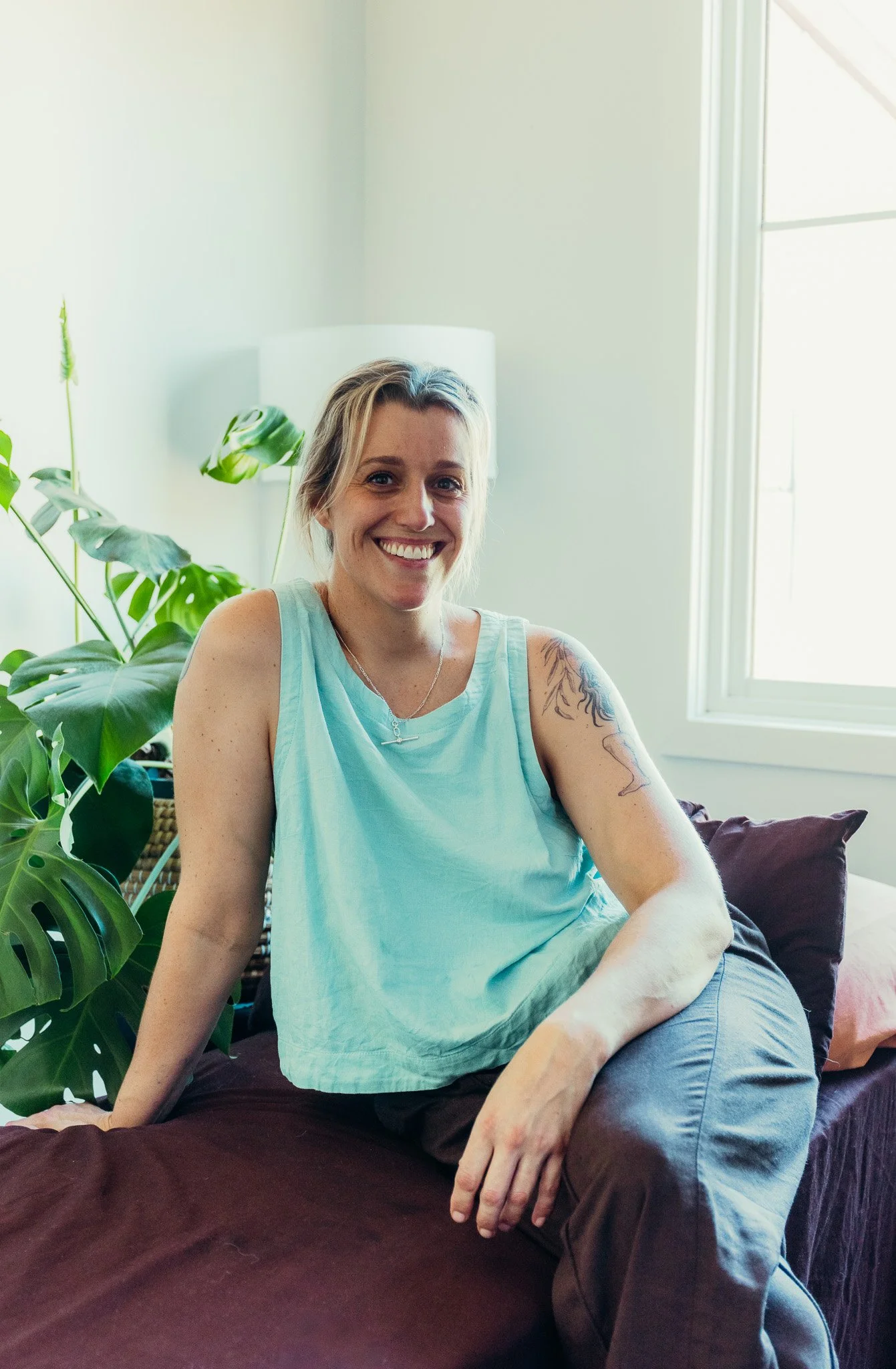 A smiling woman with blonde hair, wearing a light blue sleeveless top, sits on a dark brown couch in a bright room with a white wall, a window, and green plants.