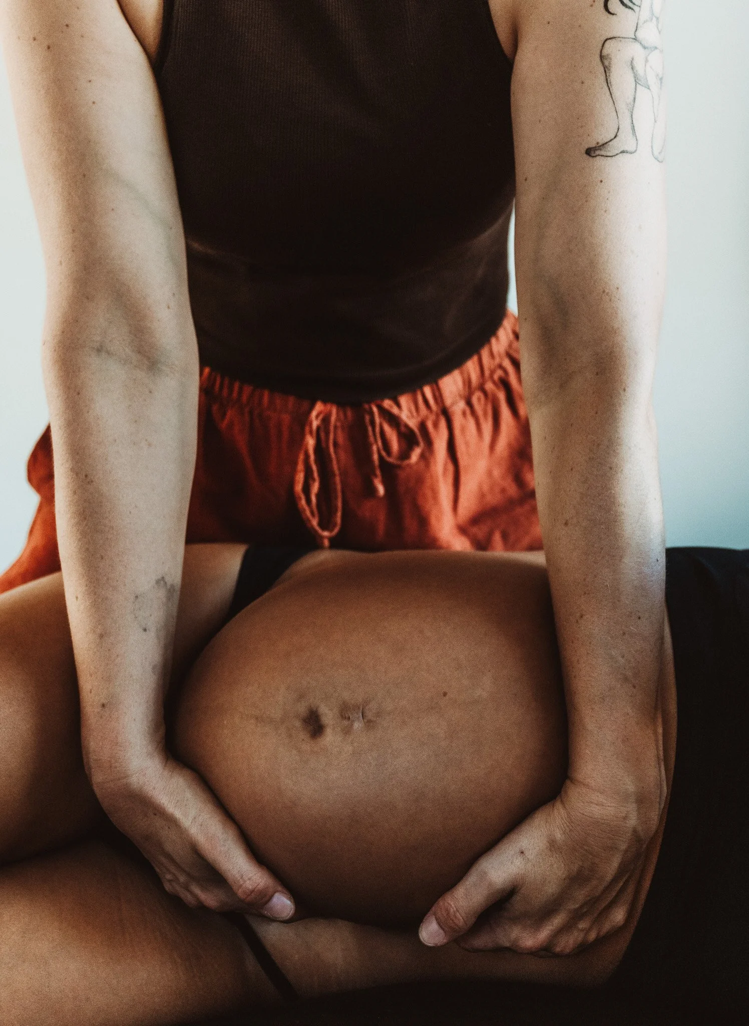 Close-up of a person's lower abdomen and hands, with another person's hands gently resting on their pregnant stomach, and part of their black shirt and orange shorts visible.