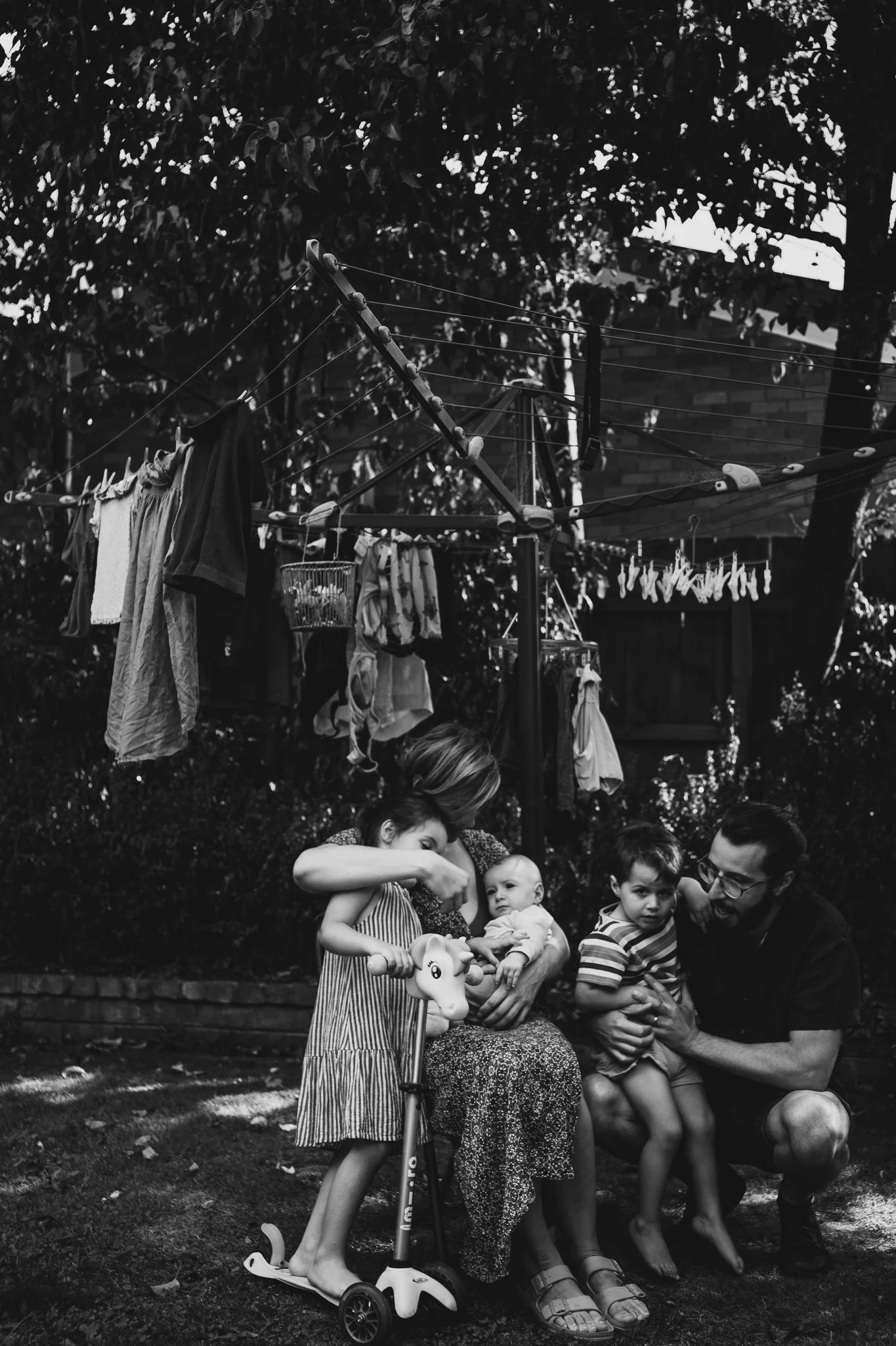 A family outdoors, including a woman with a toddler on a scooter, a young girl, a baby, a boy, and an adult man, around a bench with laundry hanging to dry in the background.