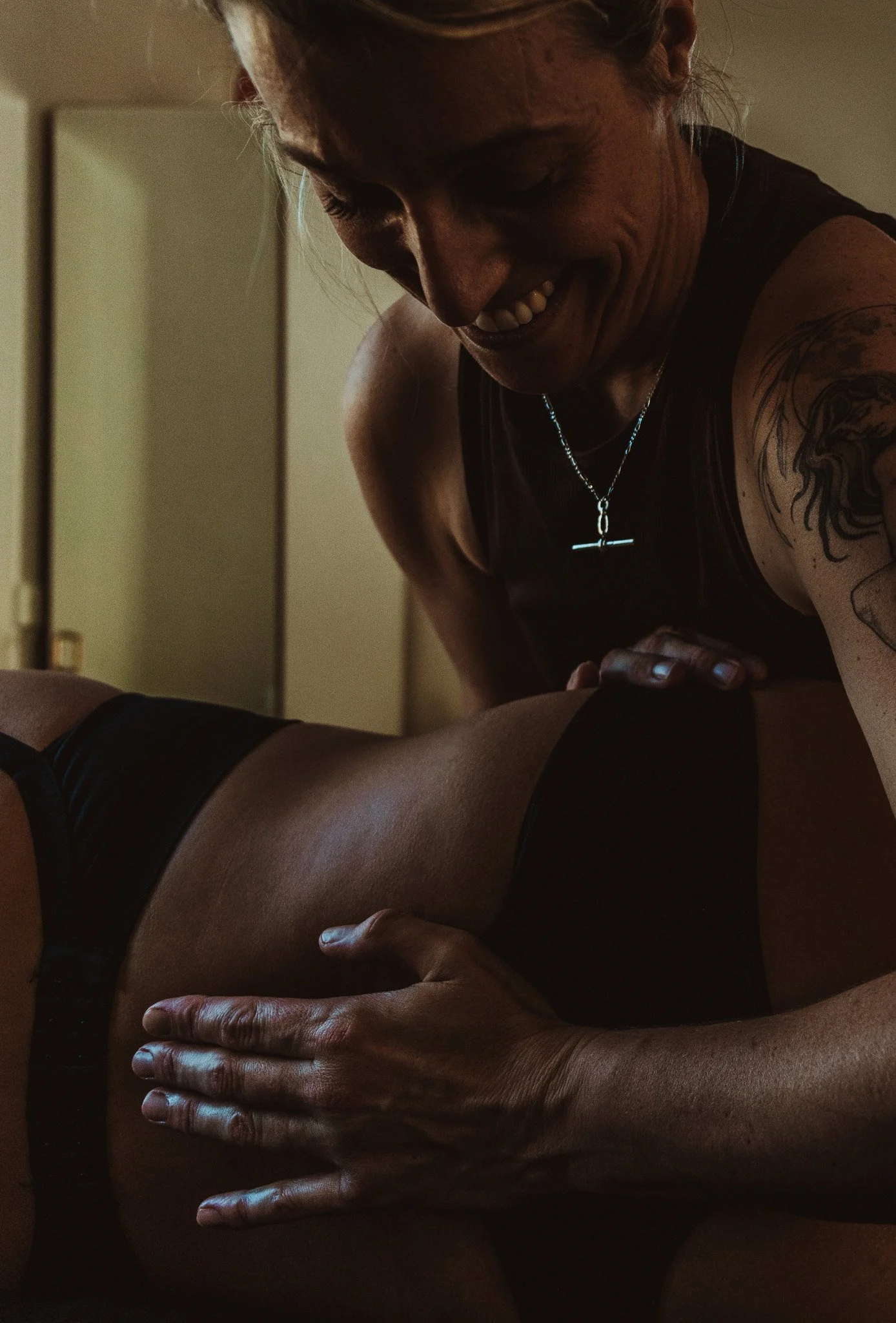 A woman with a tattoo on her arm is performing pregnancy bodywork. The woman is smiling, wearing a black tank top, a necklace with a t-bar and has her hair tied back. The setting appears to be a dimly lit room.