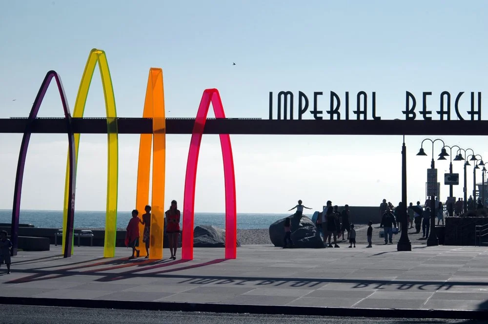 Colorful surfboard arches and Imperial Beach sign beside the oceanfront promenade.