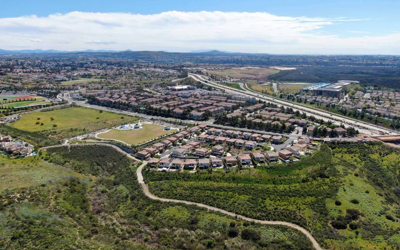 Aerial view of a suburban neighborhood with houses, a park, and surrounding green hills and open land, with a highway in the distance.