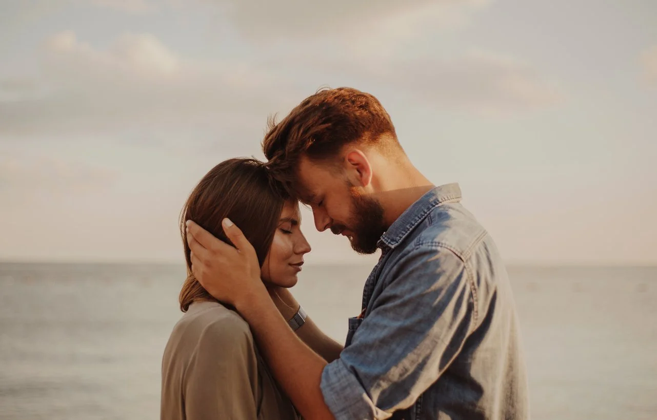 A man and woman embracing on the beach with their foreheads touching, eyes closed, during sunset.