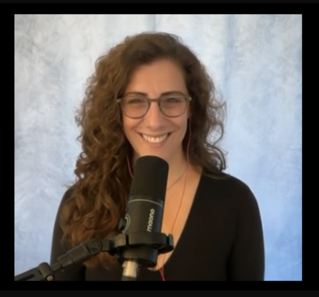 A woman with curly brown hair and glasses smiling at a microphone in front of a light blue background.