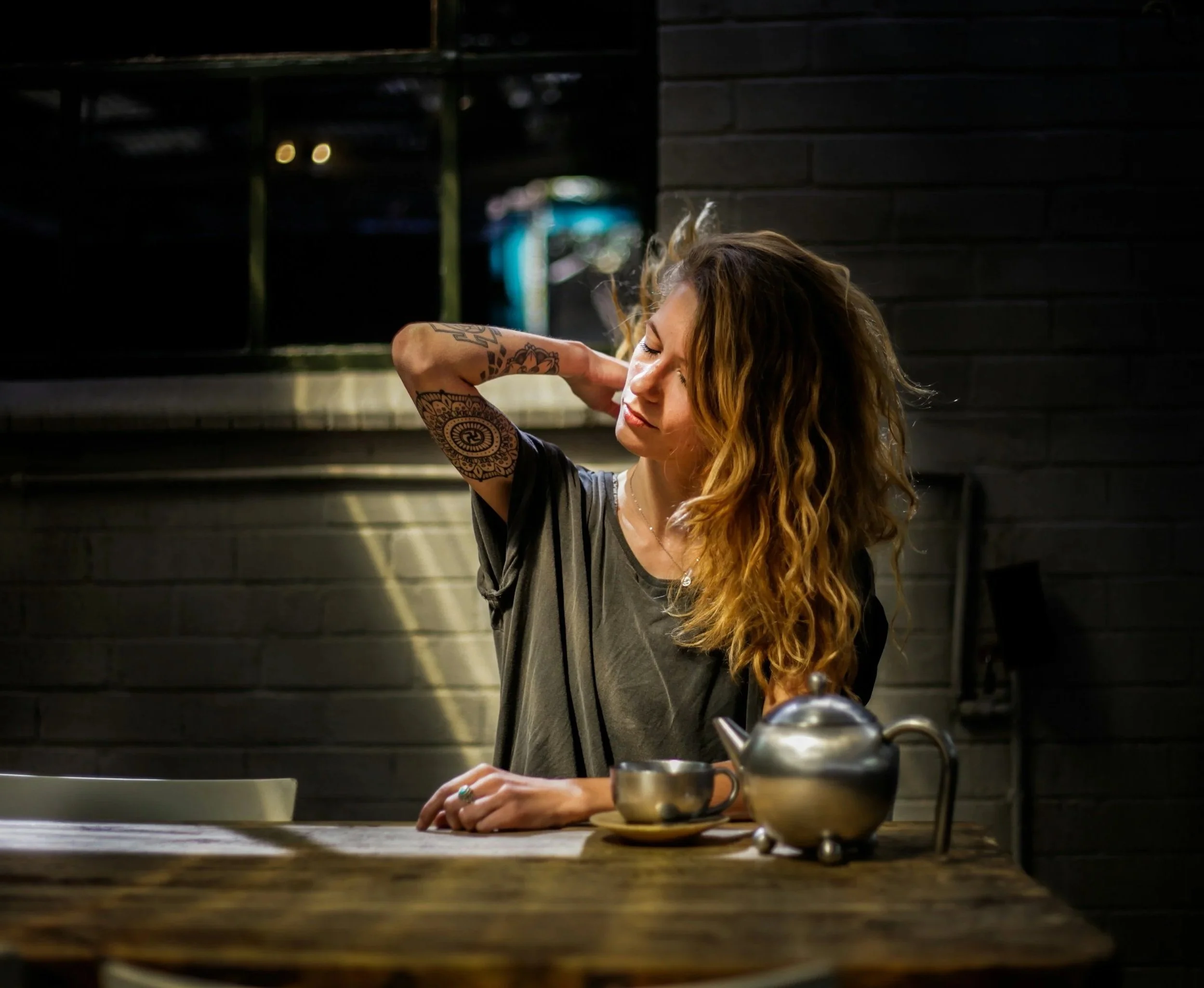 Woman with curly hair and tattoos stretching her arm behind her head, sitting at a wooden table with a teapot and cup, in a dimly lit room with a dark brick wall background.