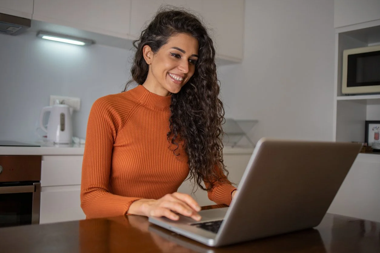 A woman with long curly hair smiling while using a laptop in her kitchen.