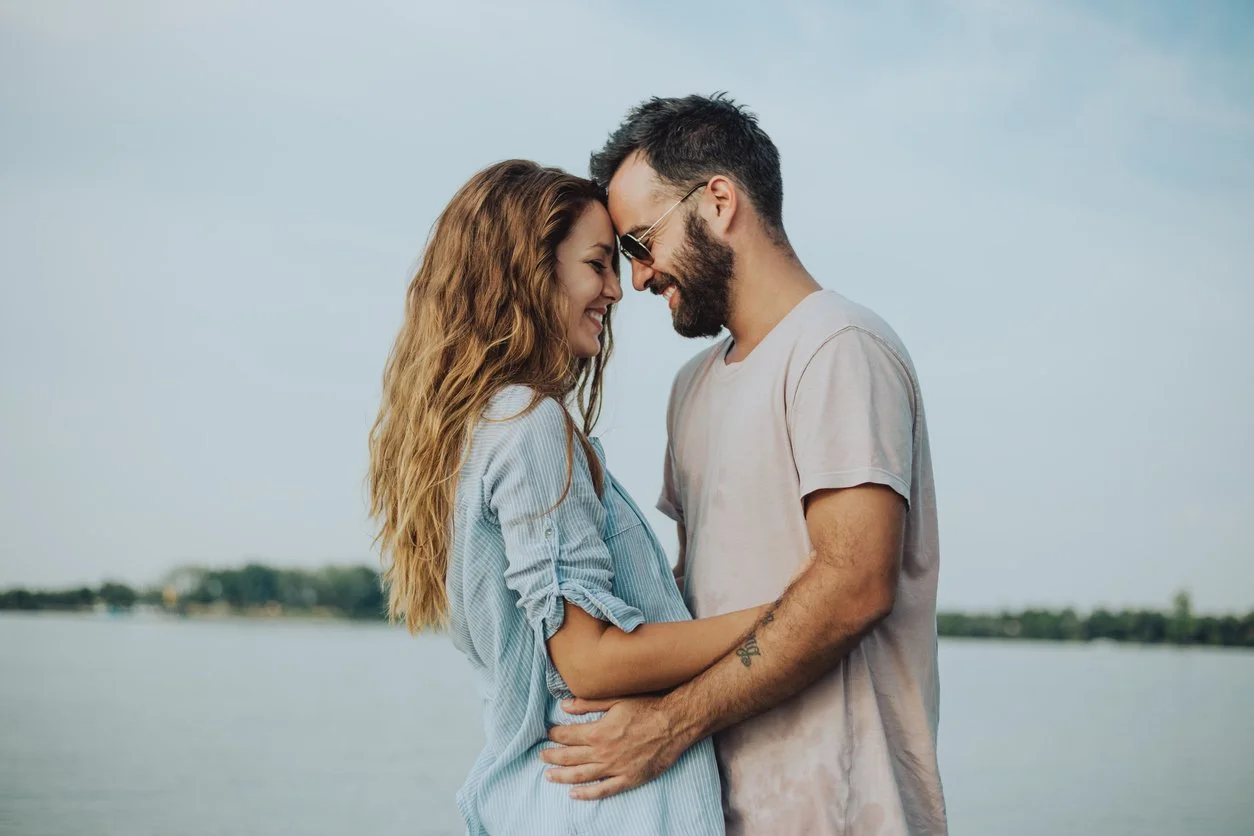 A happy couple with foreheads touching and smiling, standing outdoors near water, with a blurred natural background.