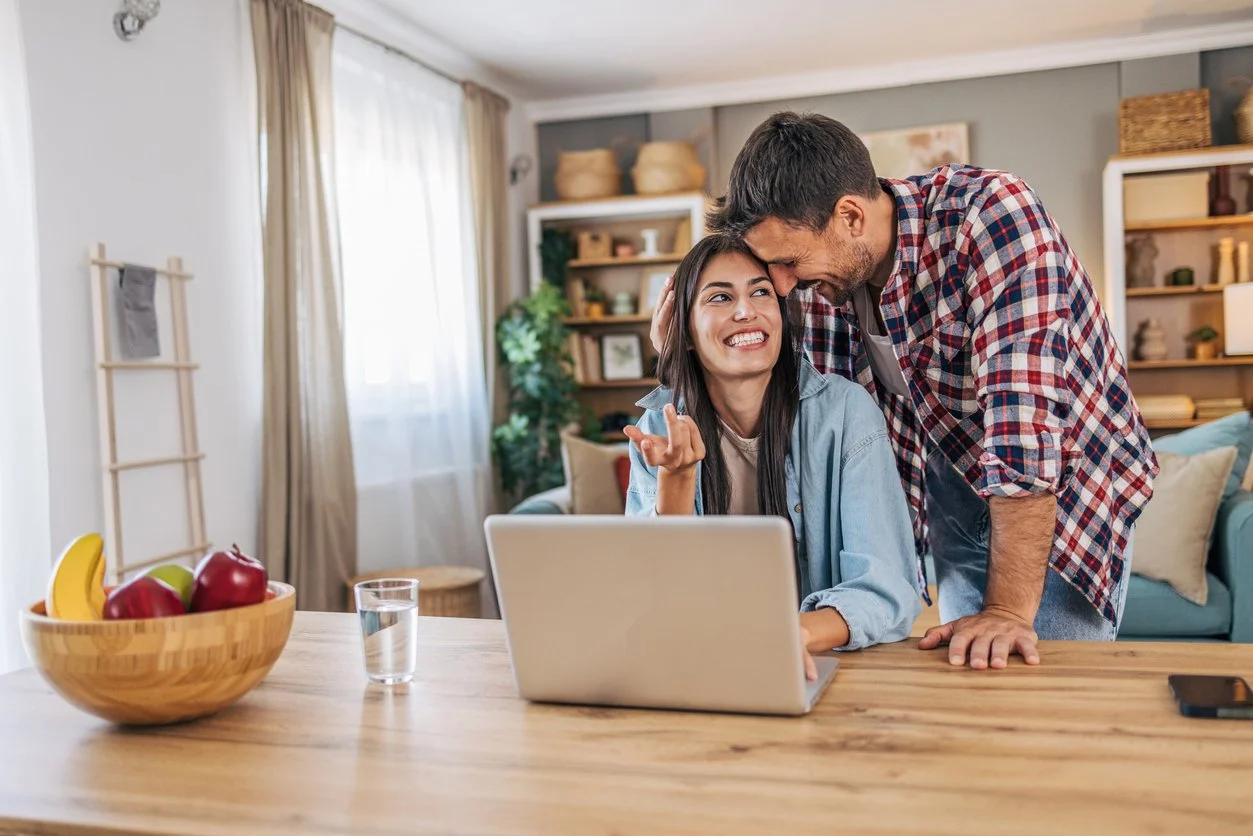 A man and woman laughing and smiling in a cozy living room, with the man leaning on the table and the woman sitting in front of a laptop, a bowl of fruit and a glass of water on the table.