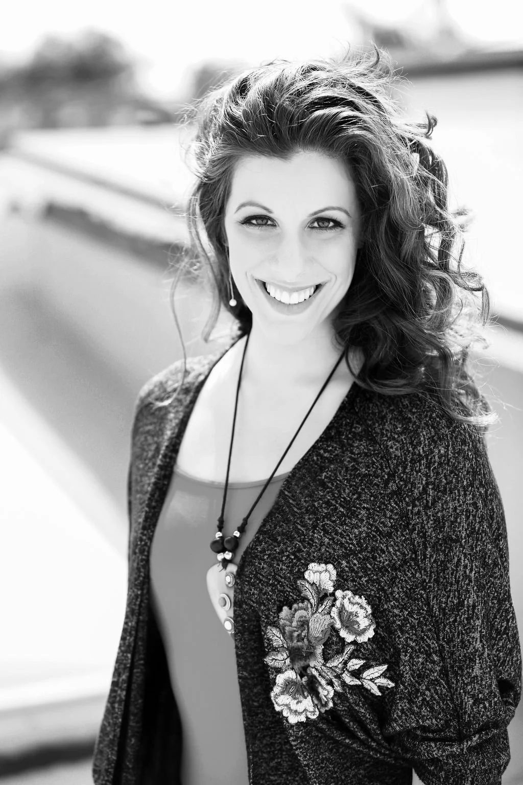 A smiling woman with curly hair wearing a dark jacket with floral embroidery and a necklace, outdoors in black and white.