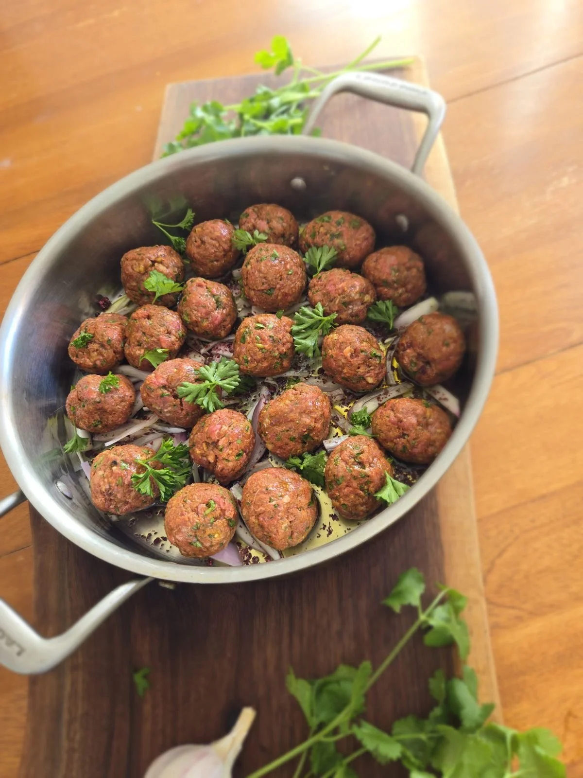 A metal pot filled with cooked meatballs garnished with fresh parsley, placed on a wooden surface with some herbs and a garlic clove nearby.