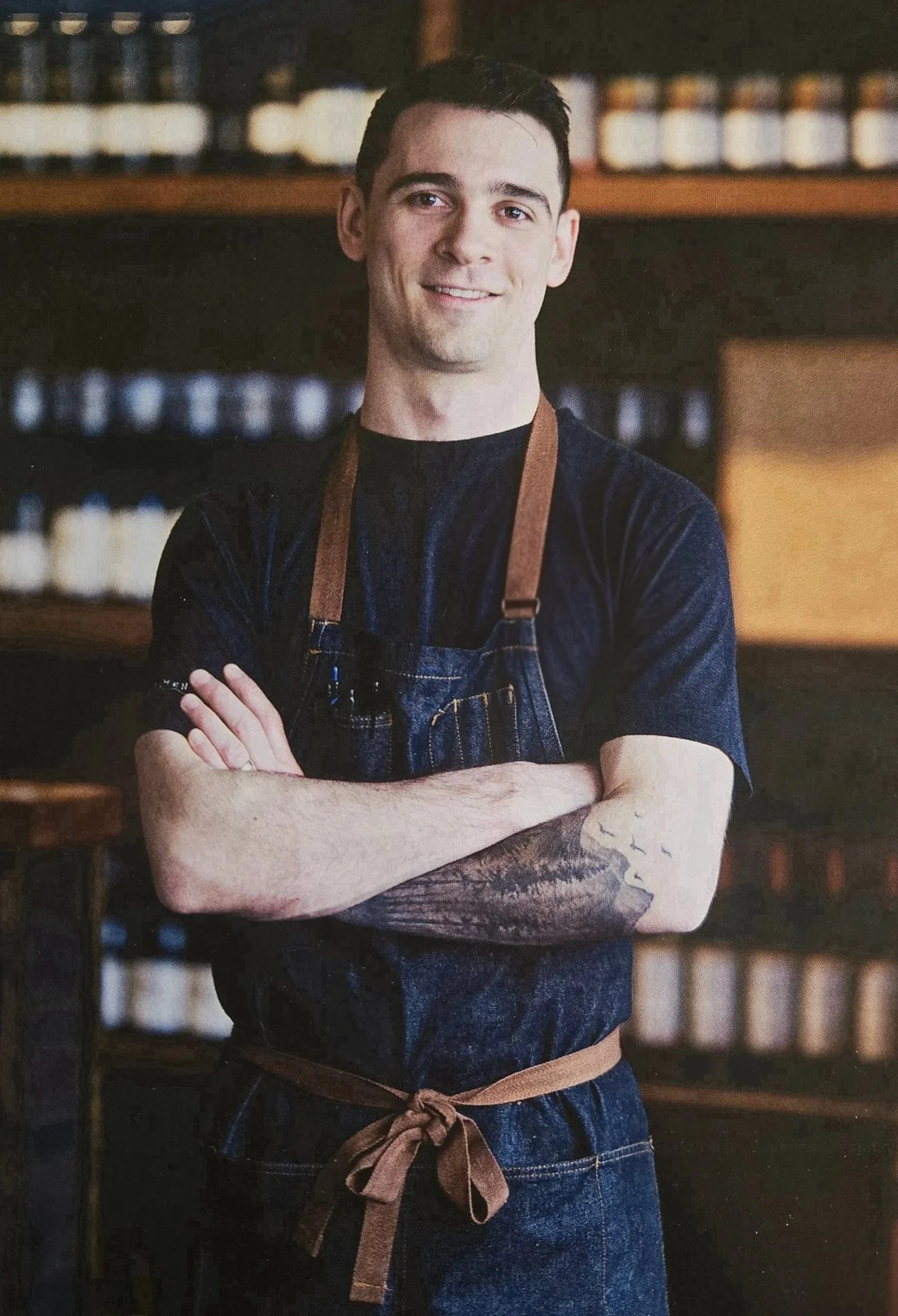 halifax meal delivery | Chef Tristan, smiling with arms crossed, wearing a black t-shirt, denim apron with brown straps and a tied brown waist apron, standing in a cozy restaurant