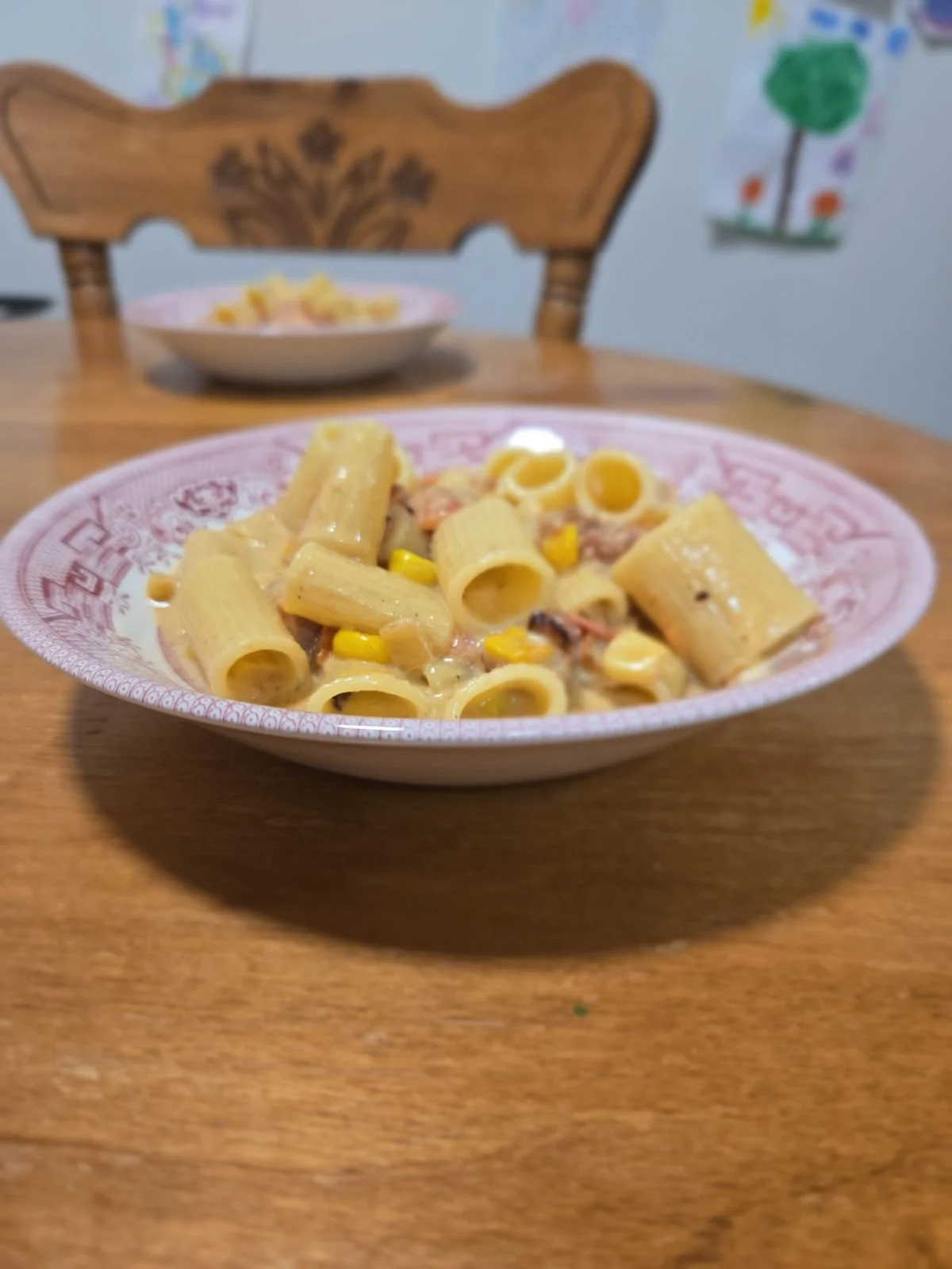 A bowl of pasta with rigatoni, corn, and creamy sauce on a wooden table. In the background, another bowl of pasta and a wooden chair are visible.