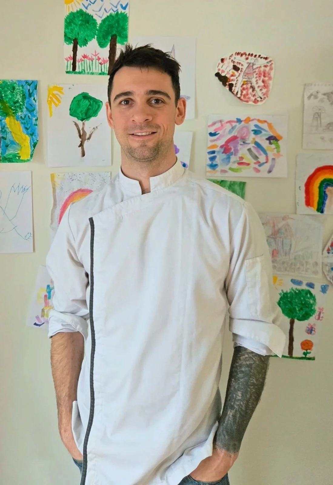 A man dressed as a chef standing in front of a wall decorated with children's colorful drawings of trees, rainbows, and abstract art.