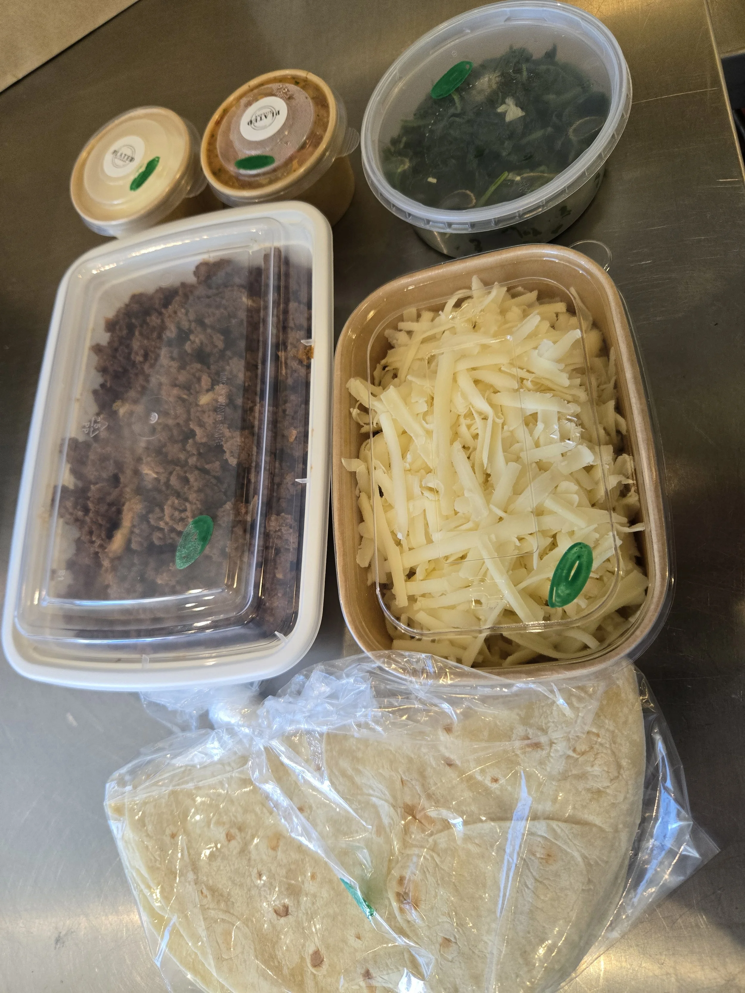 Various containers of raw and cooked ingredients, including shredded cheese, ground beef, leafy greens, and sauces on a metal surface.