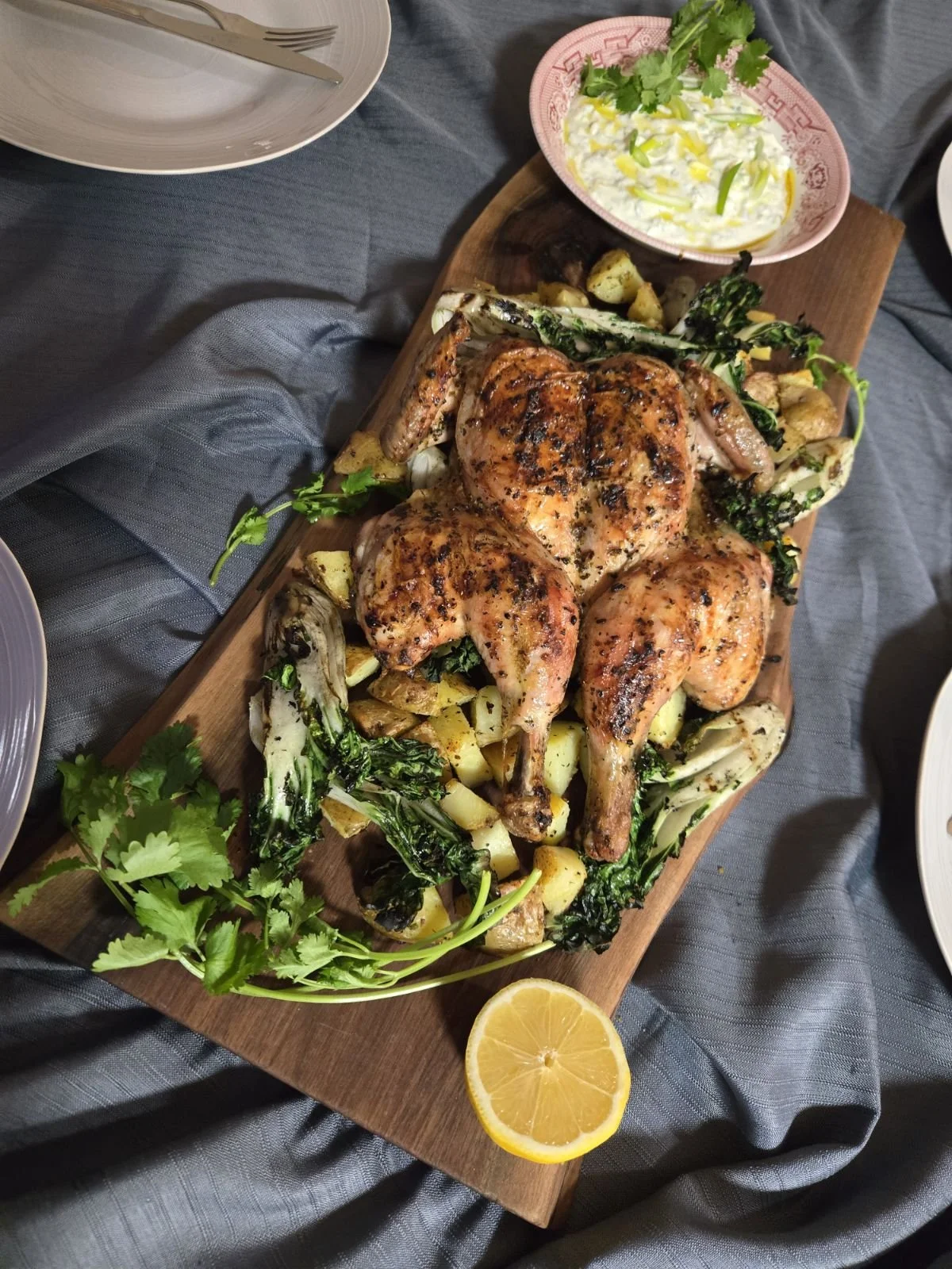 Roast whole chicken on a wooden platter with roasted potatoes, green vegetables, a lemon wedge, and a side of tzatziki garnished with cilantro, on a dark tablecloth.