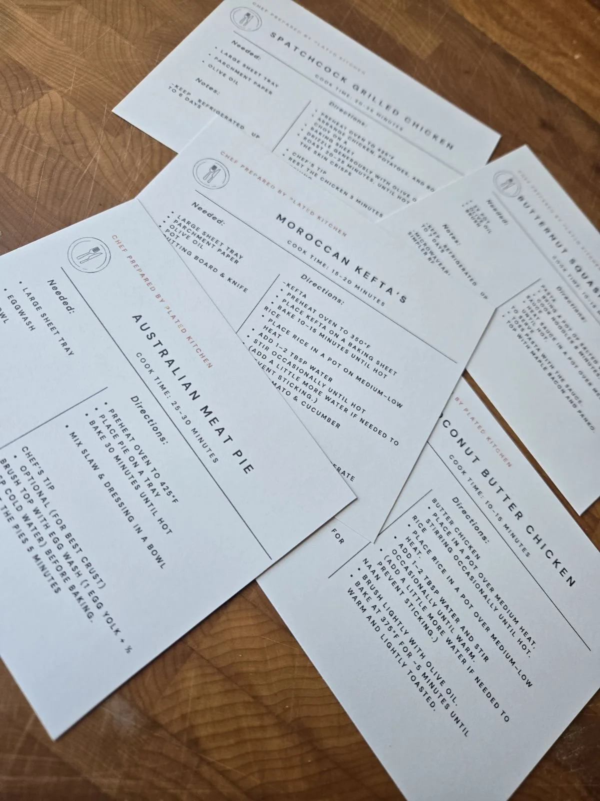 Three printed recipe cards on a wooden surface. The visible recipes are for Spatchcock Grilled Chicken, Moroccan Kefta, and Australian Meat Pie. Each card includes ingredients and cooking instructions.