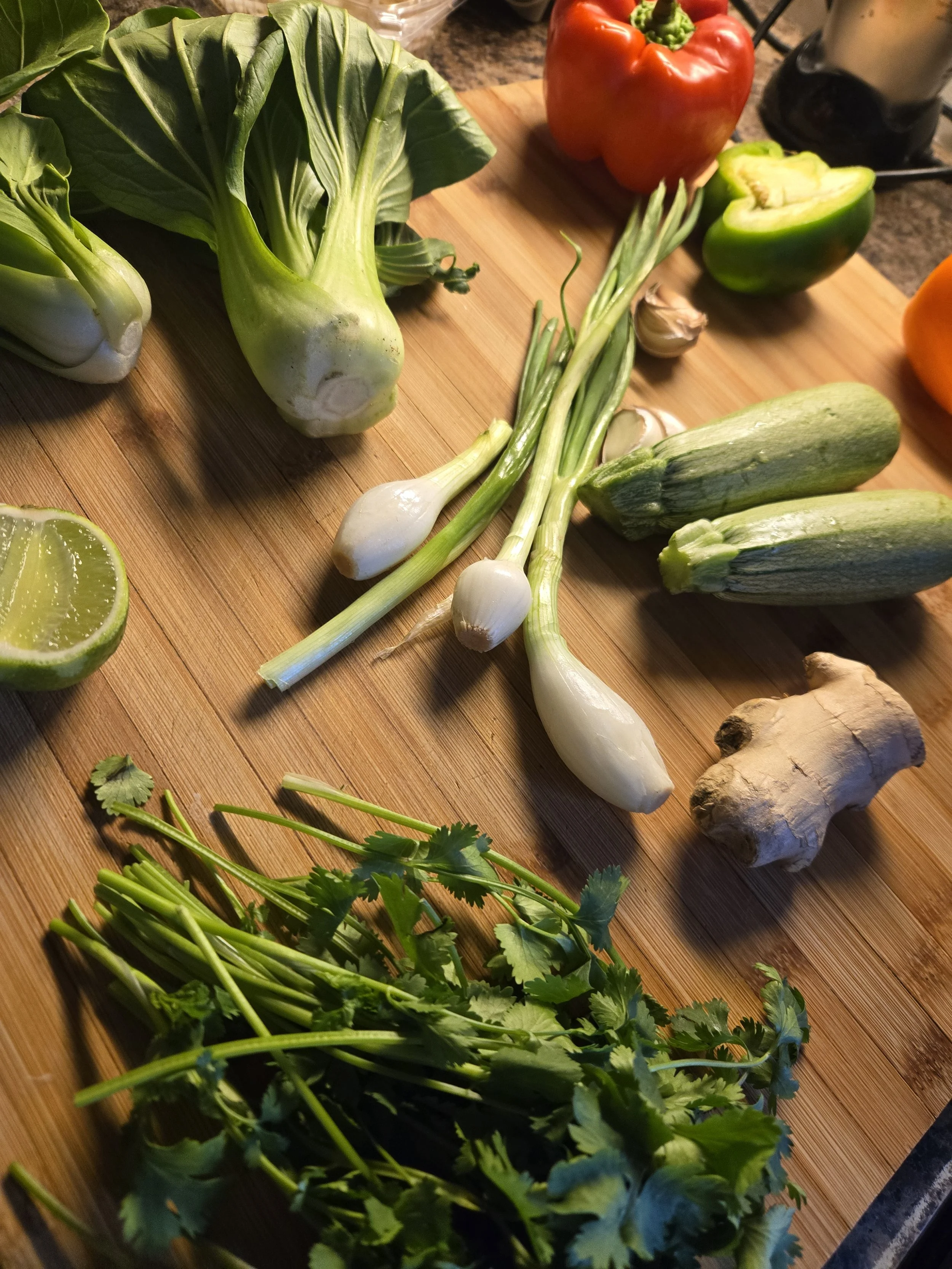 Fresh vegetables on a wooden cutting board, including a green bell pepper, a red bell pepper, green onions, garlic, ginger, lime, and cilantro.