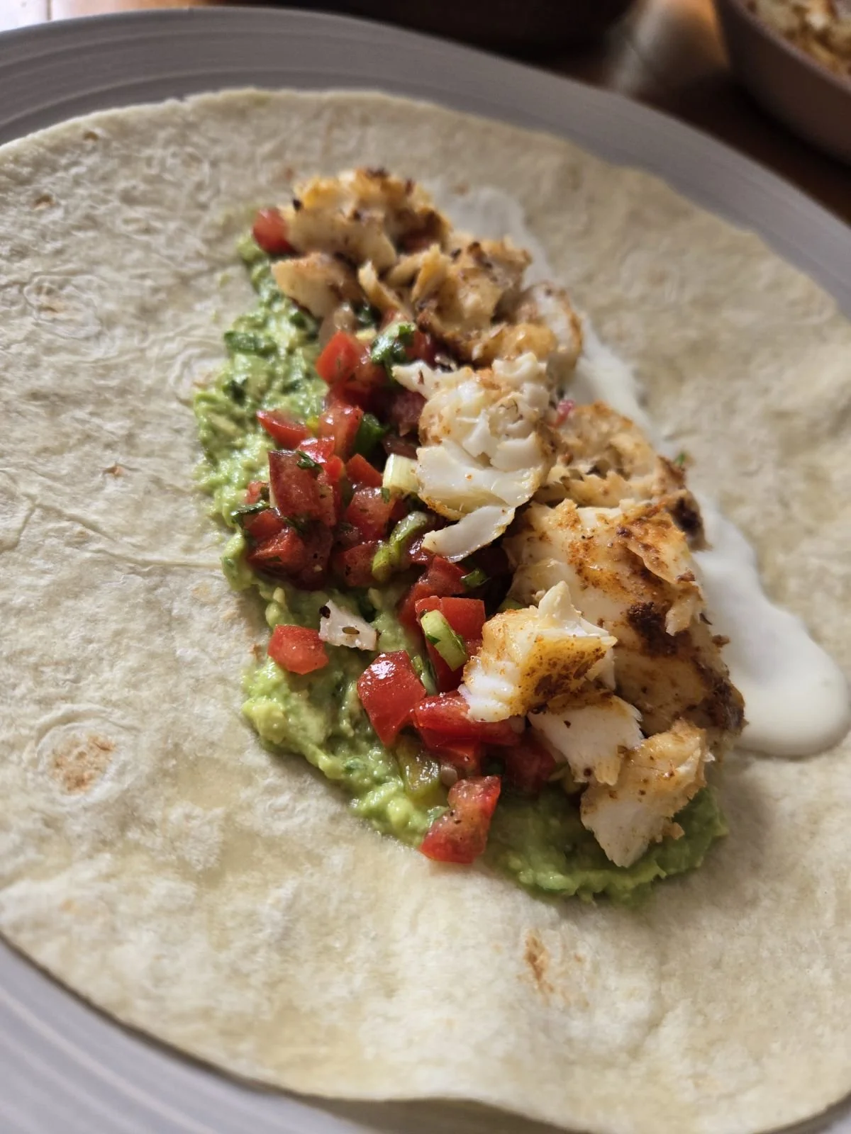 A soft taco on a plate filled with guacamole, diced tomatoes, chopped green onions, grilled chicken, and creme fraiche