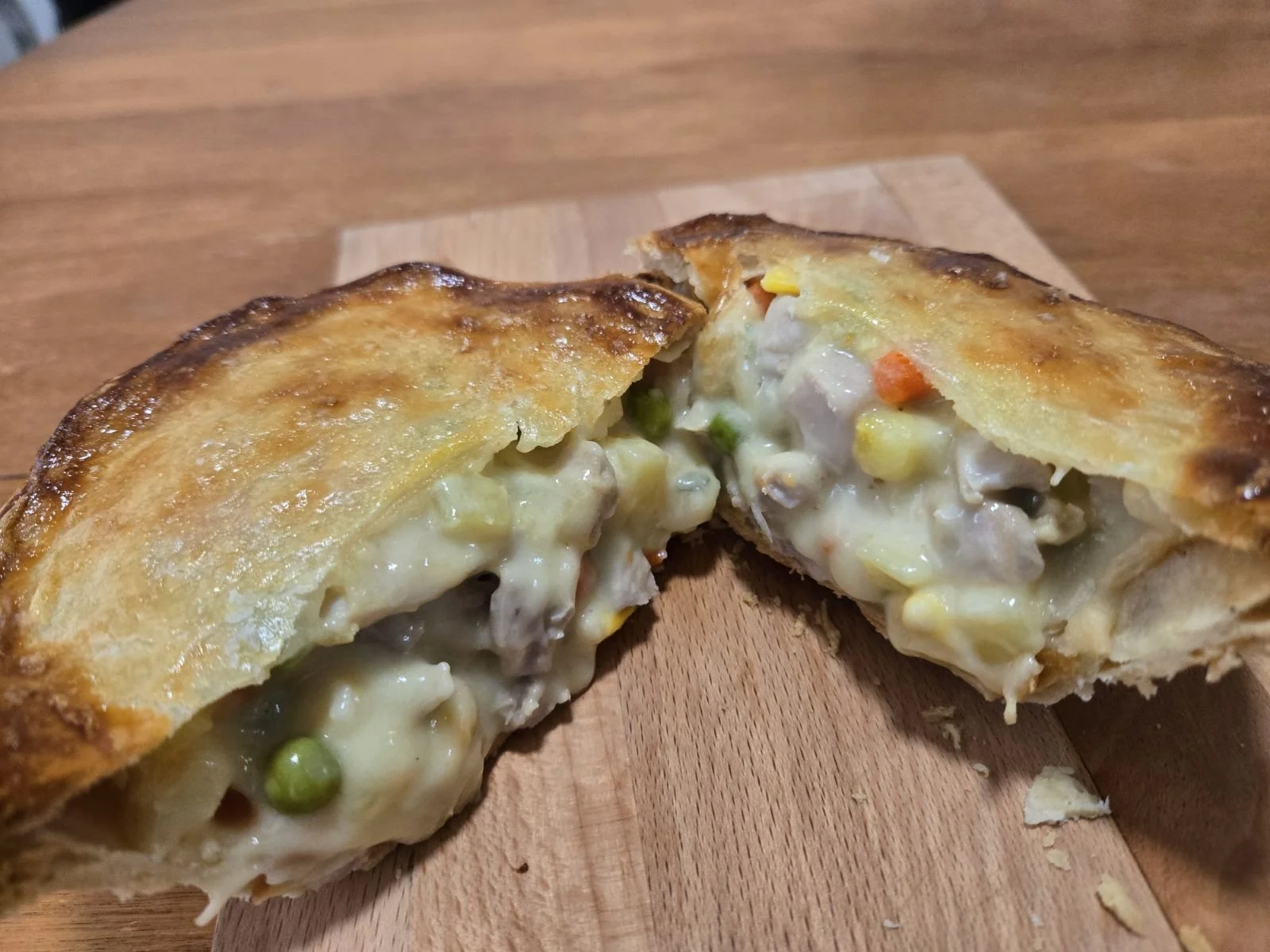 Close-up of a savory pot pie cut in half, showing a creamy filling with peas, carrots, and chunks of meat inside a flaky baked crust on a wooden cutting board.