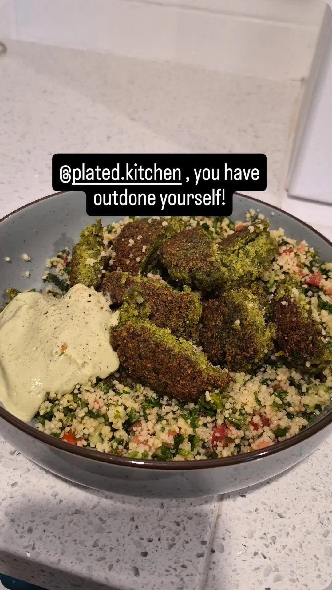 Bowl of seasoned falafel, hummus, and a bed of couscous with herbs and vegetables, on a speckled countertop.