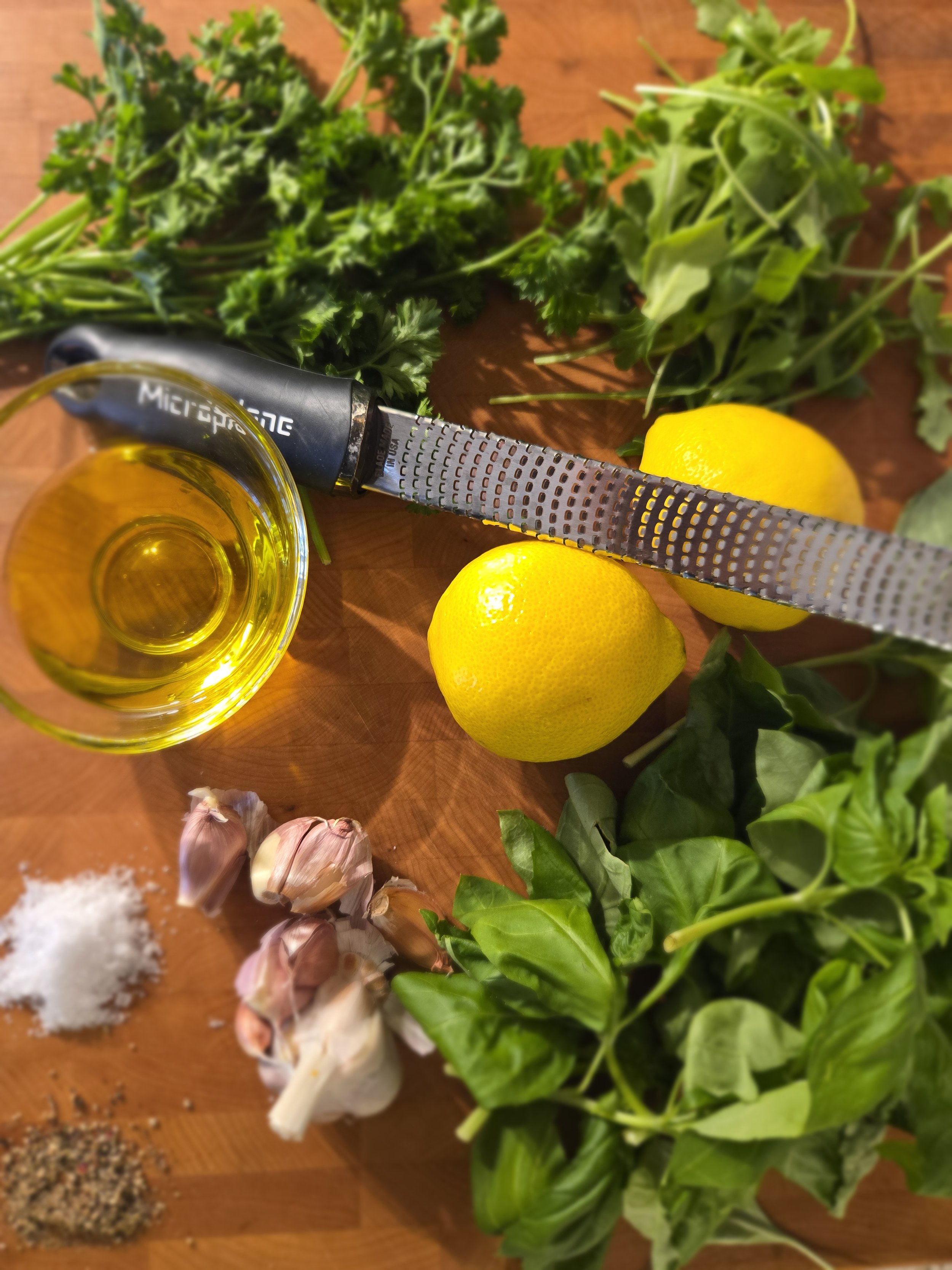 Fresh herbs including parsley, basil, and spinach, a lemon, garlic cloves, salt, freshly ground black pepper, and a garlic press on a wooden surface.
