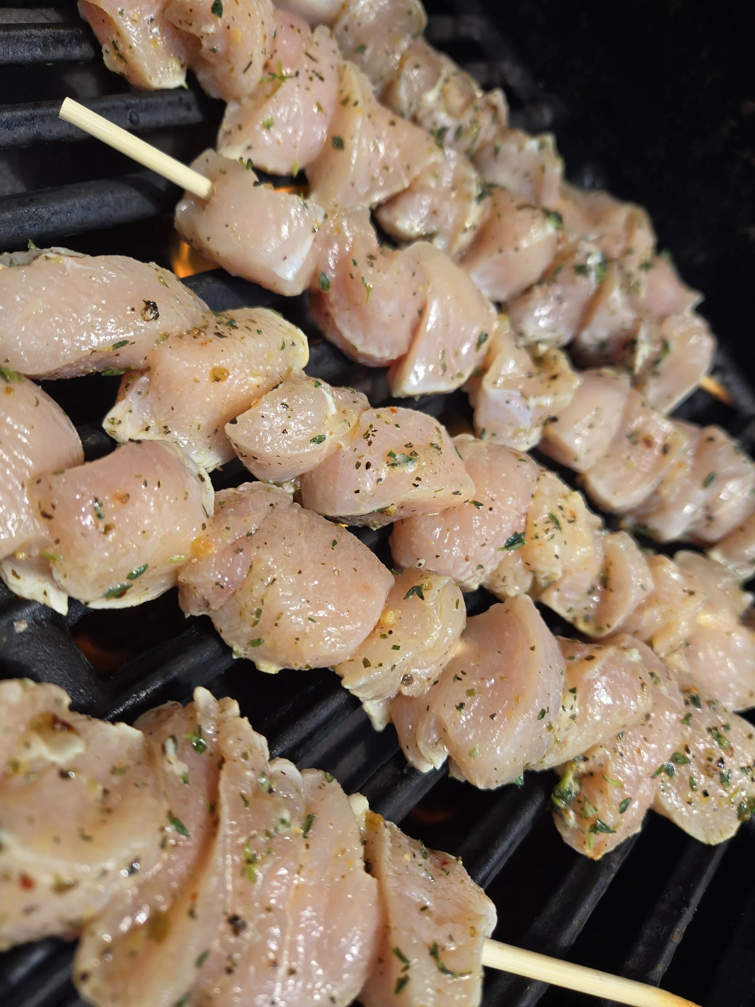Raw chicken pieces marinated with herbs and spices, threaded on skewers, cooking on a grill.