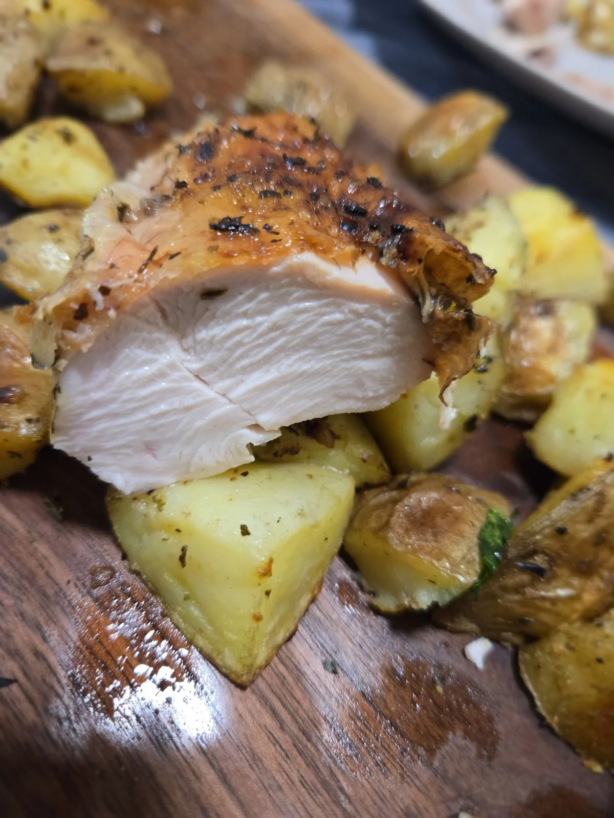 Close-up of cooked chicken breast with crispy seasoned skin, resting on a wooden cutting board with roasted potatoes around it.