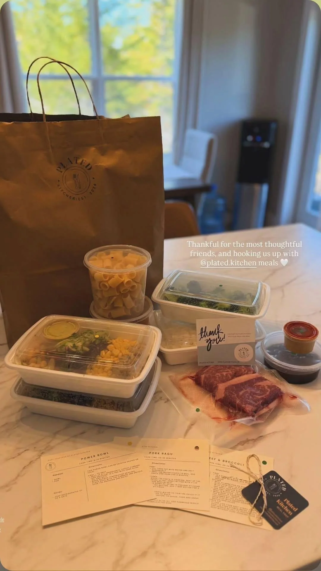 Assorted meal containers with salad, pasta, and meat, a bag from Plated Kitchen, and a thank you note on a marble table in front of a window with green trees outside.